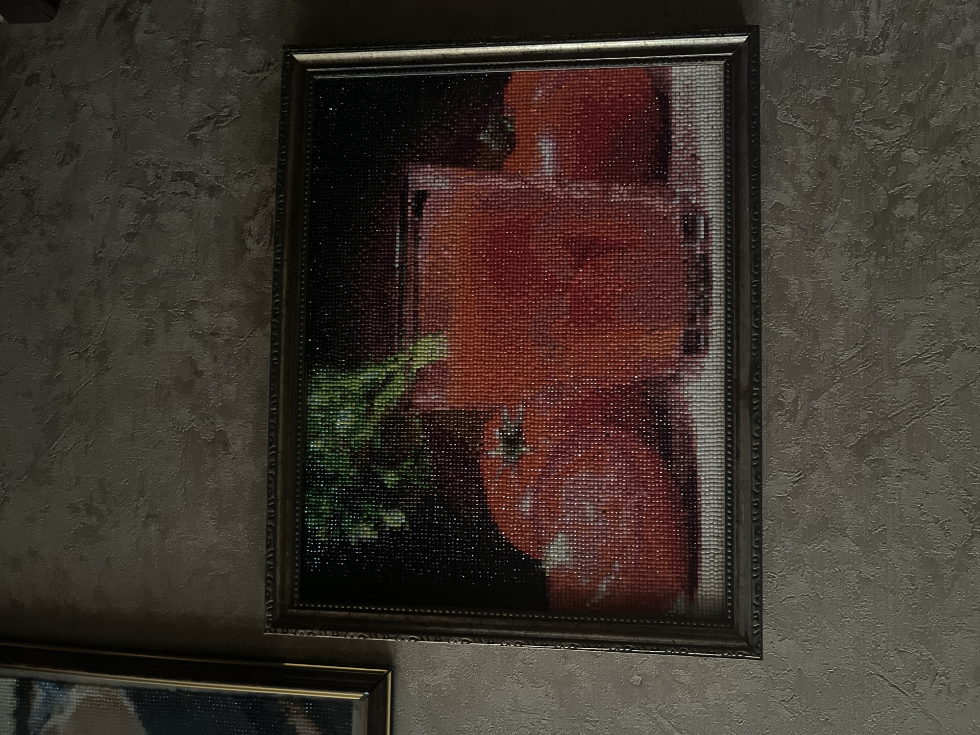 The image displays a framed diamond painting depicting a glass of tomato juice and three tomatoes, hanging on a textured gray wall. The painting is in the foreground, and another framed artwork is partially visible to its left. The lighting is dim, casting shadows and highlighting the texture of the wall and the sparkly surface of the diamond painting. The location is specified as Vladimirovka, Russia.