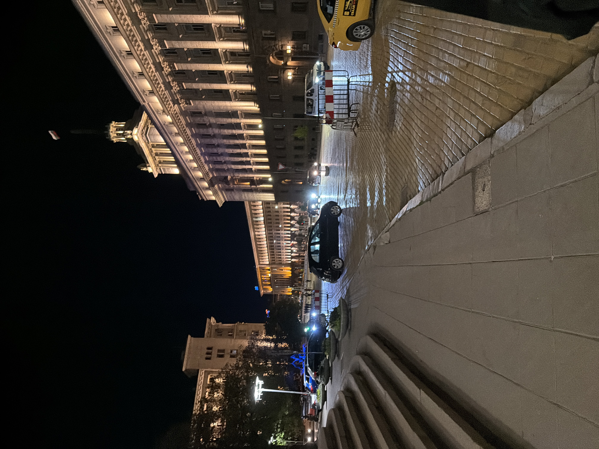 This media file depicts a city street at night, likely in Sofia, Bulgaria, after rainfall. The scene is dominated on the left by a grand, multi-story building of classical or neoclassical architecture, brightly illuminated to highlight its ornate facade and numerous windows. A flag, appearing white and red, is visible flying from a pole on top of this building.
In the mid-ground, a wet, reflective cobblestone street stretches across the frame, mirroring the strong artificial lights from streetlights and building interiors. Several vehicles are present: a yellow taxi with "TAXI" and other discernible Bulgarian text (like "OK") on its side is visible on the far right of the street. Closer to the center, a white van is partially obscured by a red and white striped barrier. A dark car is positioned further down the street, and the lights of other distant vehicles are visible.
The foreground on the right features a wide, light-colored concrete sidewalk or public plaza, with steps leading down or up. Potted plants are sparsely arranged along this pedestrian area. The sky is dark, confirming the nighttime setting. The wet surfaces and strong reflections suggest recent rain, adding to the atmospheric quality of the urban scene. No explicit human activity is clearly visible, contributing to a sense of calm in the city night.