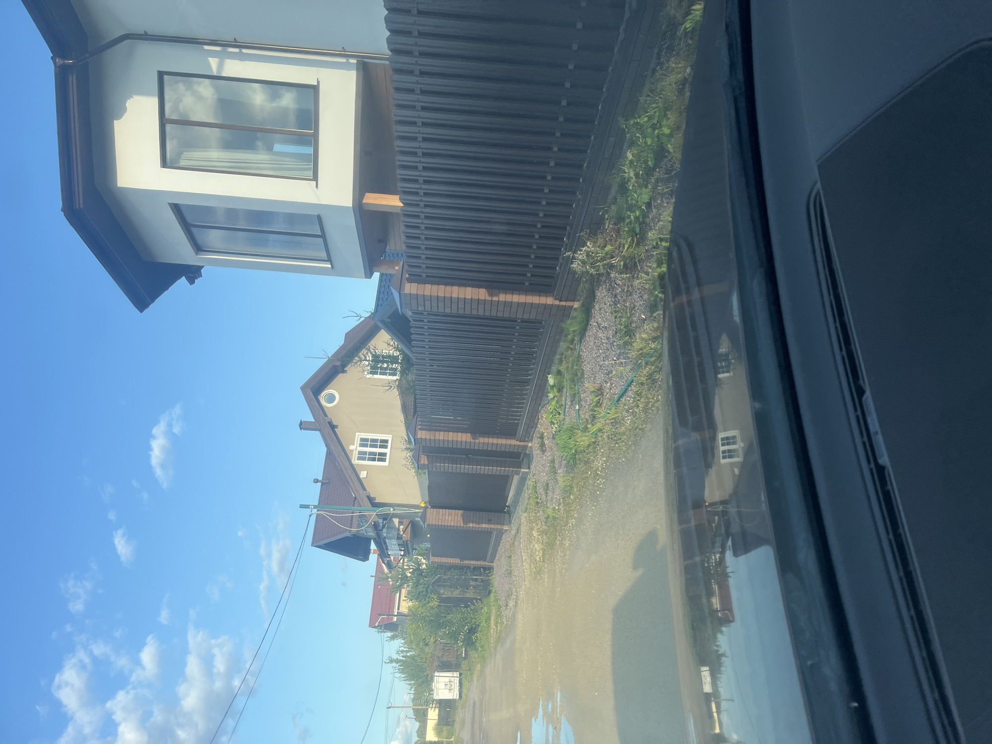The image depicts a street view in Vladimirovka, Russia, likely from the perspective of a vehicle's interior, as suggested by the reflection on the windshield and the dashboard visible in the foreground. The scene captures a residential area with houses on the right side of the road, separated by a dark-colored, vertical-slatted fence with brick pillars. The houses feature modern architecture, with one prominent house on the right having a distinctive bay window with large panes. The road surface appears to be unpaved or gravel, with puddles of water reflecting the sky and surrounding environment. Several other houses are visible further down the street, with different roof styles and colors. Vegetation, including grass and some bushes, grows along the fence line and on the side of the road. The sky is blue with scattered white clouds, indicating fair weather. No people are visible in the frame.
