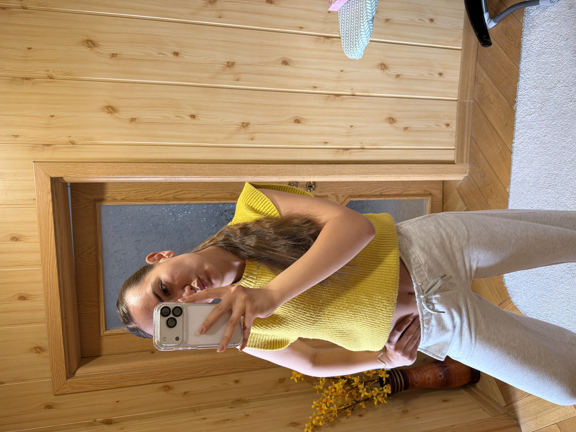 A young woman is taking a selfie using her smartphone. She is wearing a yellow knitted top and light gray sweatpants. She is standing indoors in front of a wood paneled wall with a framed door. A vase with yellow flowers is visible in the lower part of the image. The floor is wood and a light colored carpet is visible in the background. The location is likely indoors, possibly a home, but Buhovo, Bulgaria, is not evident within the frame.