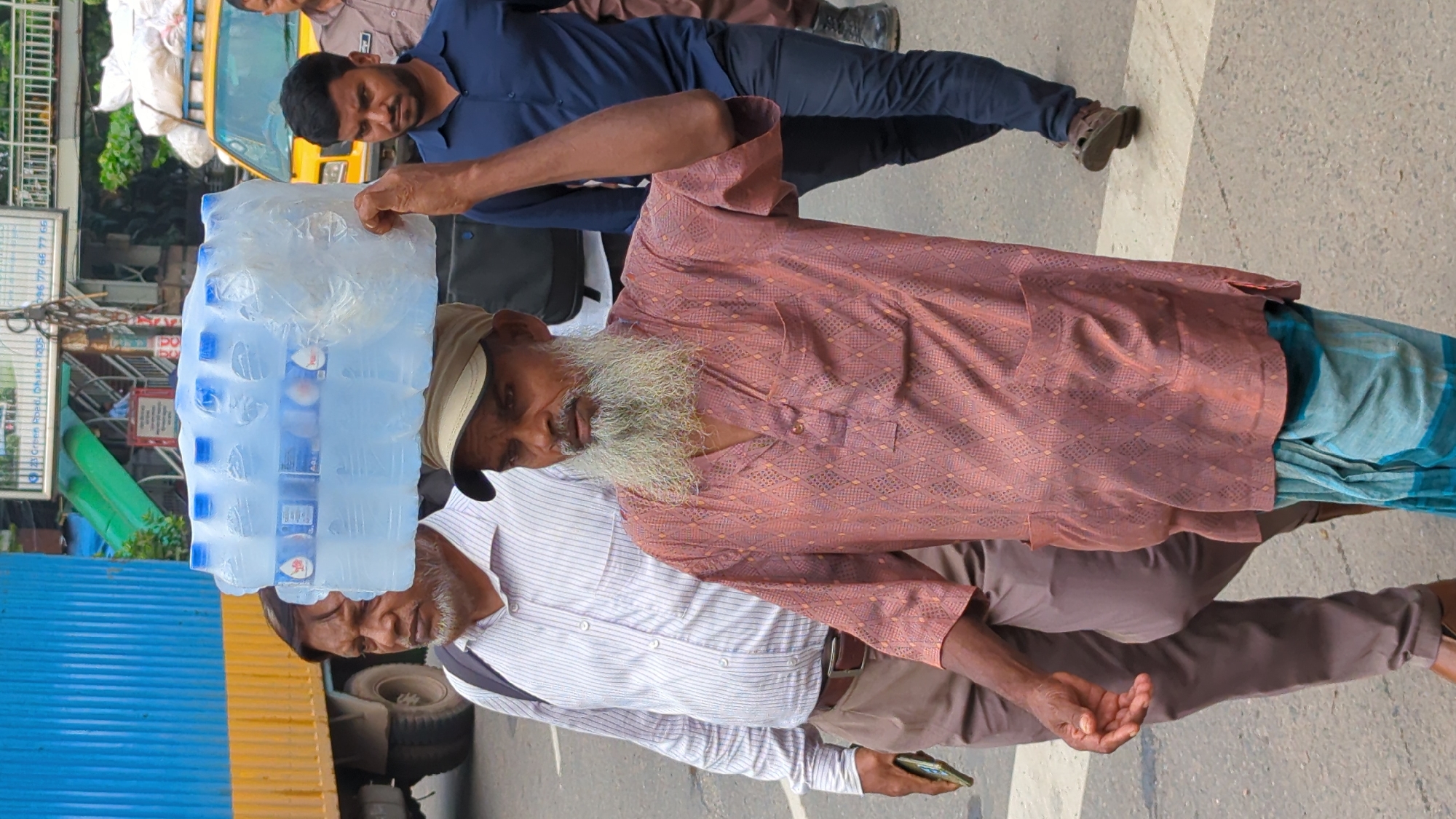 A man with a long white beard and a tan baseball cap carries a large pack of bottled water on his head. He is wearing a maroon patterned shirt and a blue and white checkered lungi. He is walking on a city street. Behind him, another man in a white striped shirt and brown pants holds a smartphone in his left hand. To the right of the man carrying water, another man in a dark blue shirt and dark pants is walking. In the background, a yellow truck is parked next to a large blue truck. The scene appears to be in Dhaka, Bangladesh, judging by the presence of typical street vendors and the style of clothing.