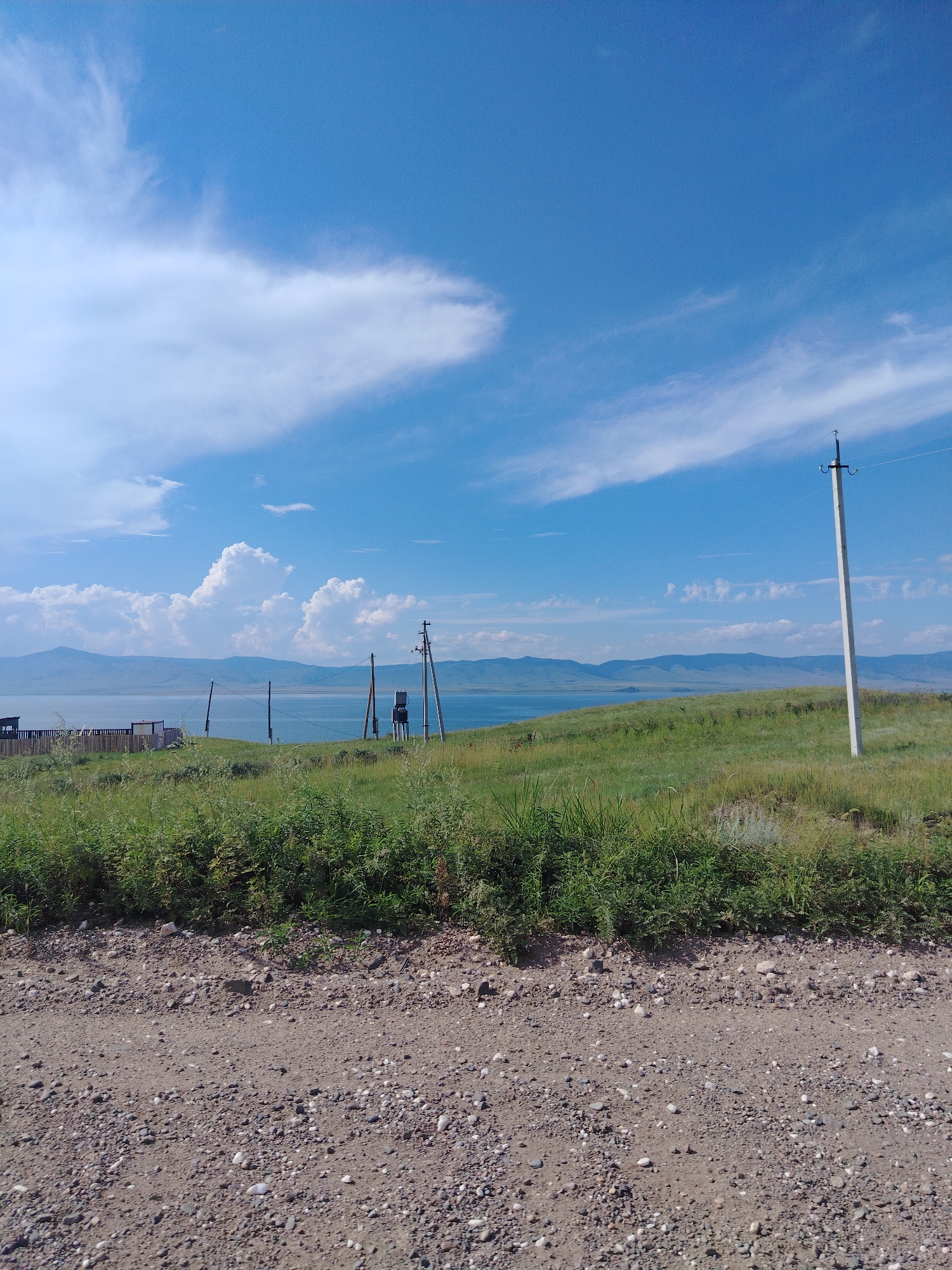 This is a medium-shot, eye-level, landscape photograph of a rural scene under a bright blue sky with scattered white clouds. In the foreground, a dirt road made of gravel and small stones stretches horizontally across the bottom of the frame. Behind the road, a grassy hill slopes upwards, covered in green vegetation with tall grasses and some smaller, lighter-colored plants.

Mid-ground, a body of water, likely a lake, is visible, bordered by a distant, rolling mountain range under the sky. On the left side of the water, a wooden fence with vertical posts and horizontal rails encloses a small area. To the right of the fence, a dark structure, possibly a small building or shed, is partially visible.

Further to the right on the hill overlooking the water, there are two utility poles. The one on the left is shorter and appears to have some equipment attached to it. The pole on the right is taller and stands prominently against the sky, with wires extending from it. The overall environment suggests a natural, undeveloped area. There are no people or animals visible in the photograph. The scene is static and depicts a serene, natural landscape. The location is identified as Tsaydam, Russia.