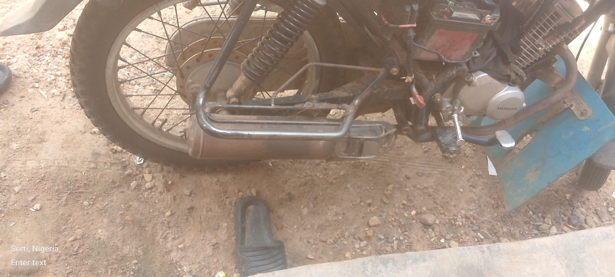 A close-up, eye-level, slightly angled outdoor shot focuses on the rear wheel and exhaust of a motorcycle, resting on a dirt and gravel surface. The motorcycle's frame, shock absorber, and a portion of the engine with the "HONDA" logo are visible. A blue fender is also present. A black sandal is lying on the ground near the rear wheel. The ground is a mixture of soil, small stones, and debris. The text "Serti, Nigeria," and "Enter text" appear in the lower-left corner of the image.