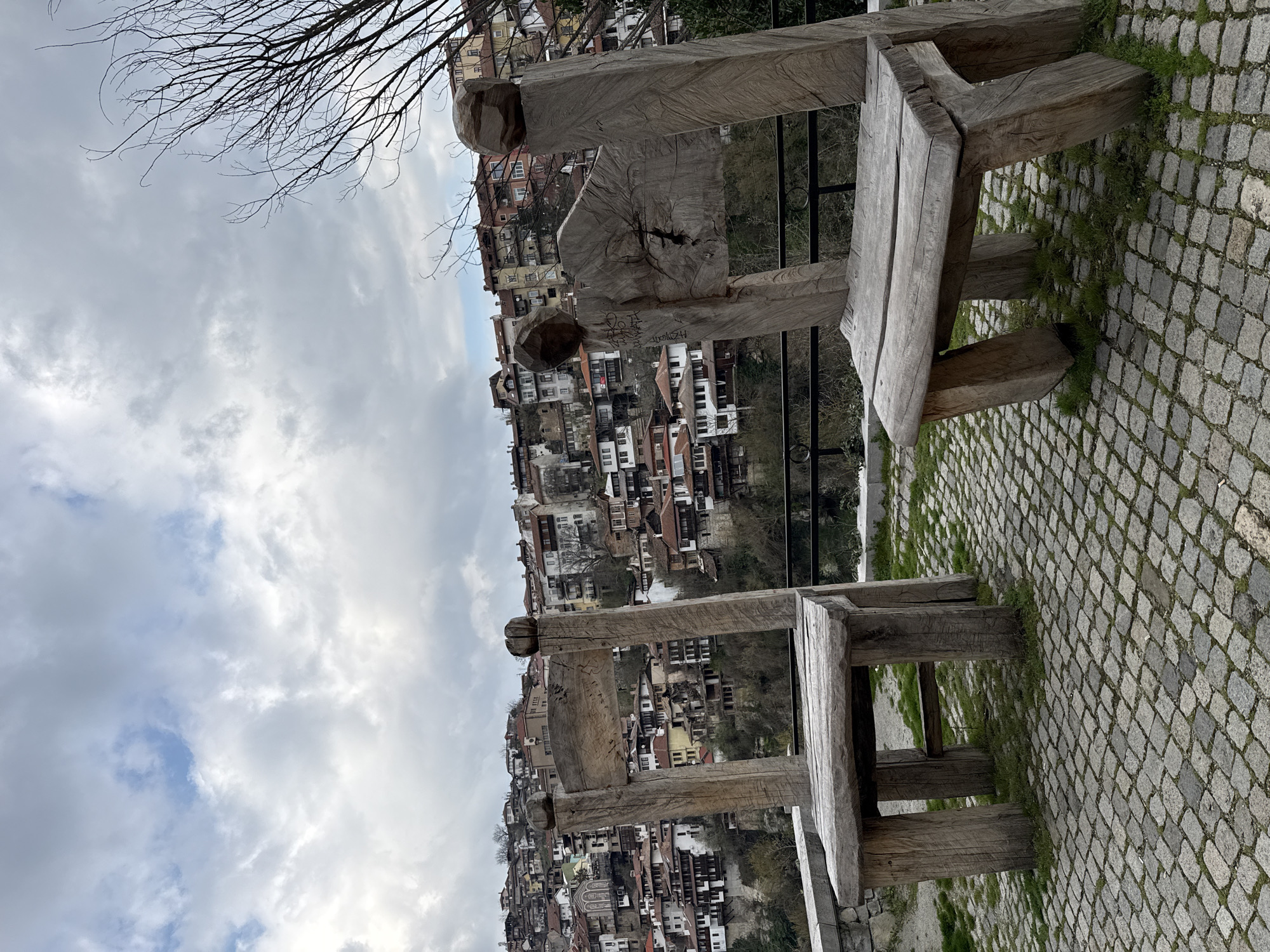 Here is a detailed description of the image:
The image shows two wooden benches overlooking a hillside town. The benches are rustic in style, appearing to be made from roughly hewn logs. They are placed on a paved area with small cobblestones and patches of grass.
Behind the benches, a densely packed town rises up the hillside. The buildings are a mix of white and brown, with red-tiled roofs. The architecture suggests an old, possibly historic, European or Middle Eastern settlement.
The sky is overcast with a mix of grey and white clouds. A few bare branches of a tree are visible in the top left corner.
The location is in the city of Unknown, Unknown.