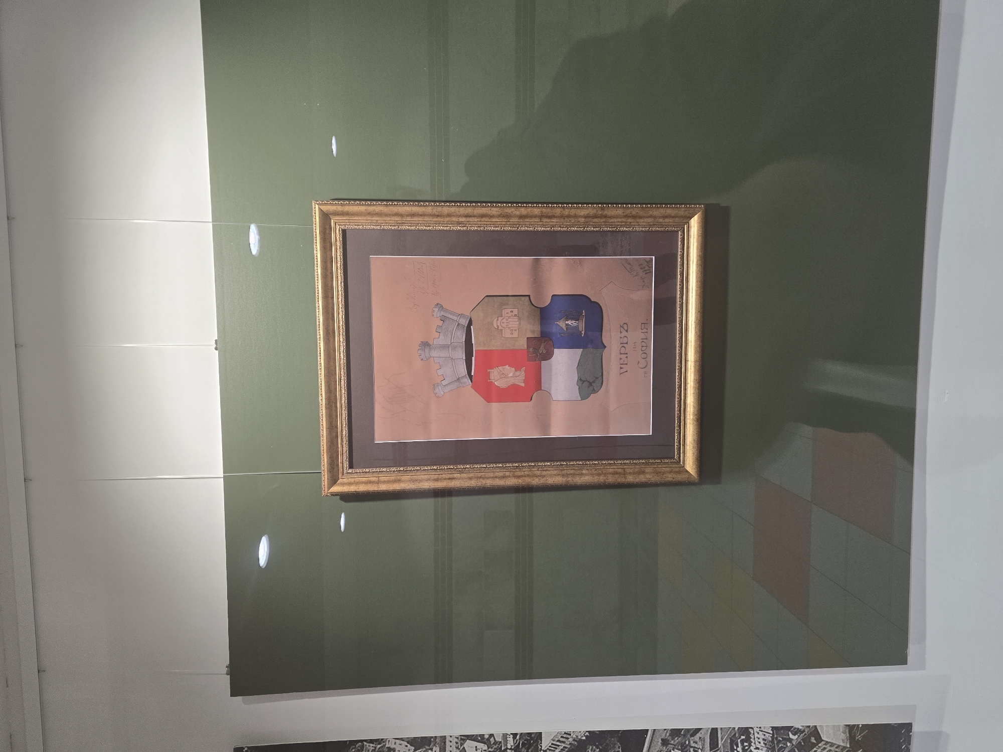 A framed drawing of the coat of arms of Sofia, Bulgaria hangs on a green wall. The drawing is in a gold-colored frame. The drawing itself is of a shield divided into quarters, with a fortress tower on the left. The text on the drawing includes the name "Sofia". The drawing is mounted on a brown background. The wall is green with some vertical stripes. The lighting in the room is bright. Part of another display case is visible at the bottom of the frame.