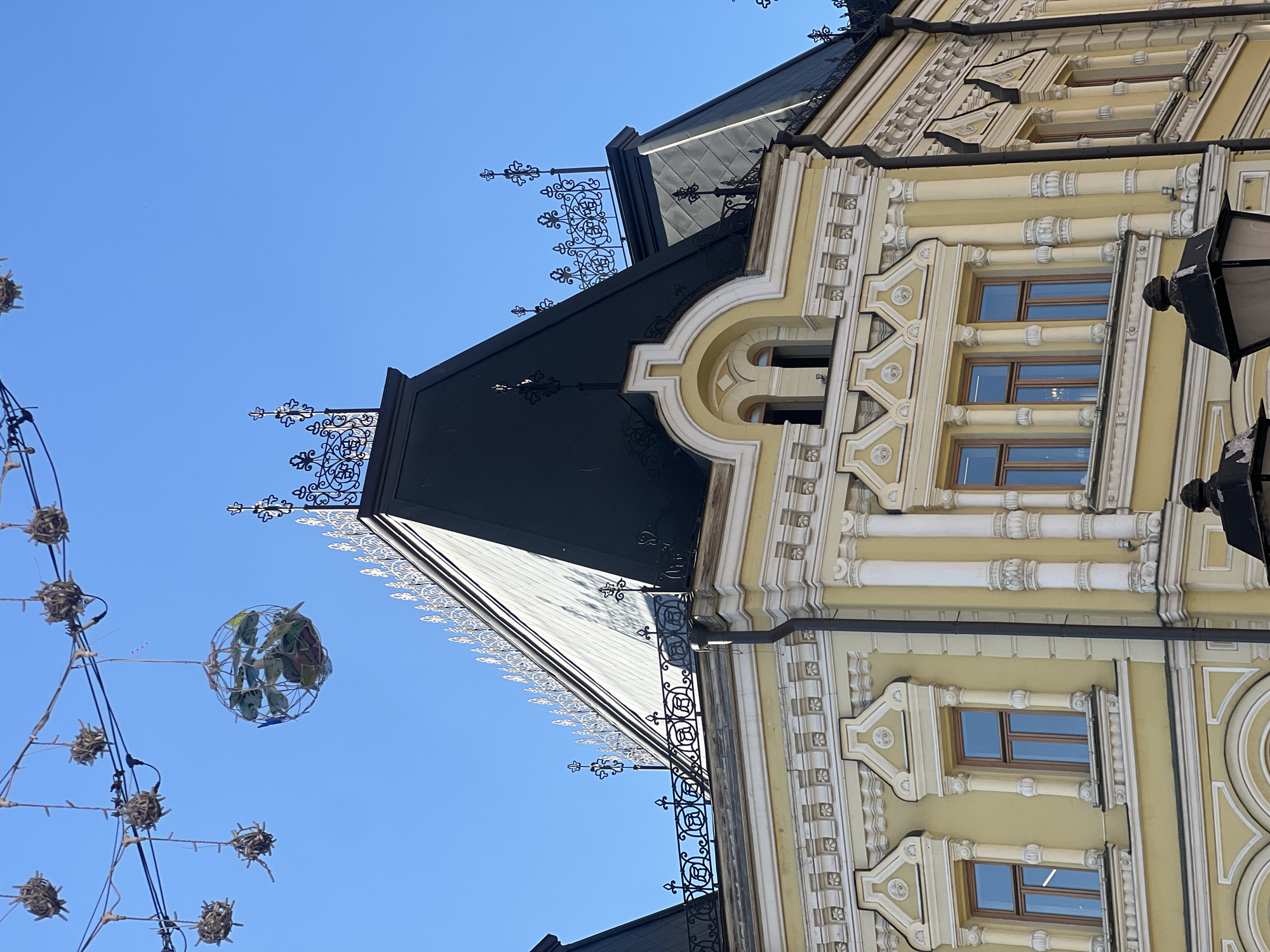 The media file shows a close-up, angled view of a building facade against a clear blue sky, suggesting a daytime scene with fair weather. The building exhibits ornate architectural details, painted in a pale yellow with white decorative elements. These include elaborate window frames, decorative columns, and moldings. The roof is dark, possibly slate or metal, with decorative ironwork along the eaves and peaks.
On the left side of the image, string lights are visible, decorated with small, spherical ornaments that appear to be made of natural materials like twigs or straw. A larger, spherical object, also made of wire and containing some greenery, hangs from a string light, suggesting an outdoor decoration, possibly for a holiday or festive occasion.
The camera angle is tilted, and the image appears to be rotated, making it difficult to definitively identify specific architectural styles without further context, but the ornamentation suggests a historical or neoclassical design. No people are visible in the frame. The overall impression is one of urban architecture and decorative elements against a bright sky.