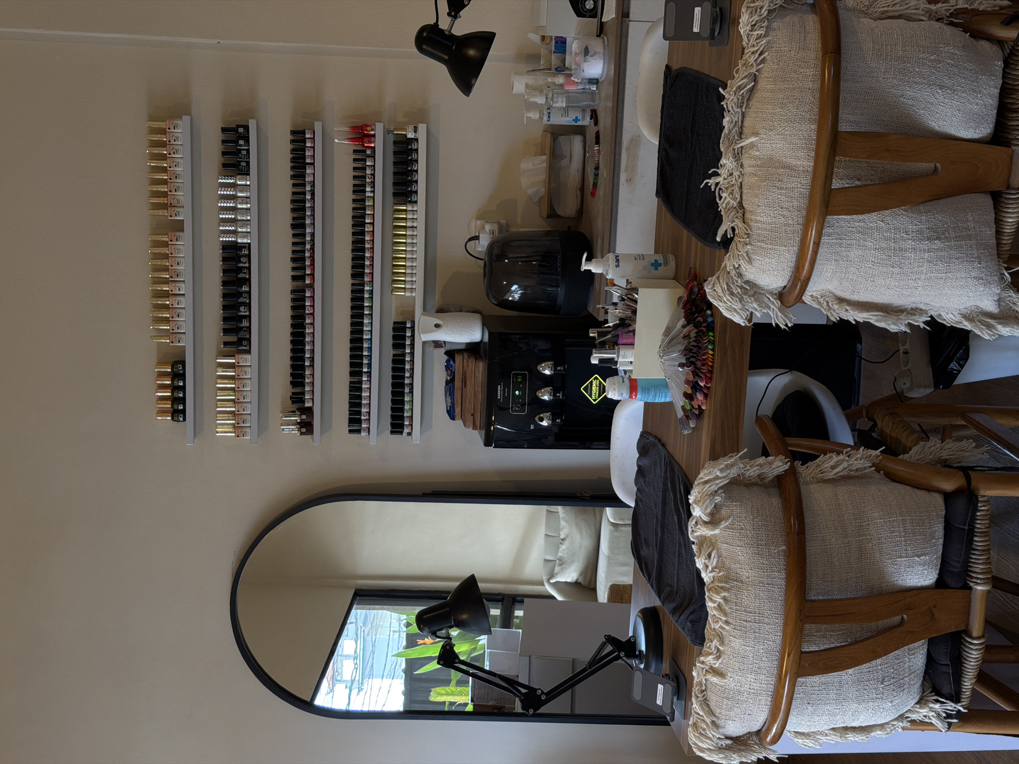 The image shows a clean and organized nail salon station in North Kuta, Indonesia. The station features a wooden table with two chairs, both adorned with tasseled cream-colored cushions. On the table, there's a black desk lamp, a black towel, and a display of nail polish color swatches. To the right of the table, a water dispenser is visible, along with a tissue box and various bottles of nail care products and hand sanitizers. The wall behind the station is lined with shelves displaying a wide selection of nail polish bottles in various colors and finishes. A large arched mirror reflects the room, showing a glimpse of a comfortable seating area with a cream-colored sofa. The overall impression is one of a professional and well-equipped nail salon ready to provide services. There are no people visible in the image.