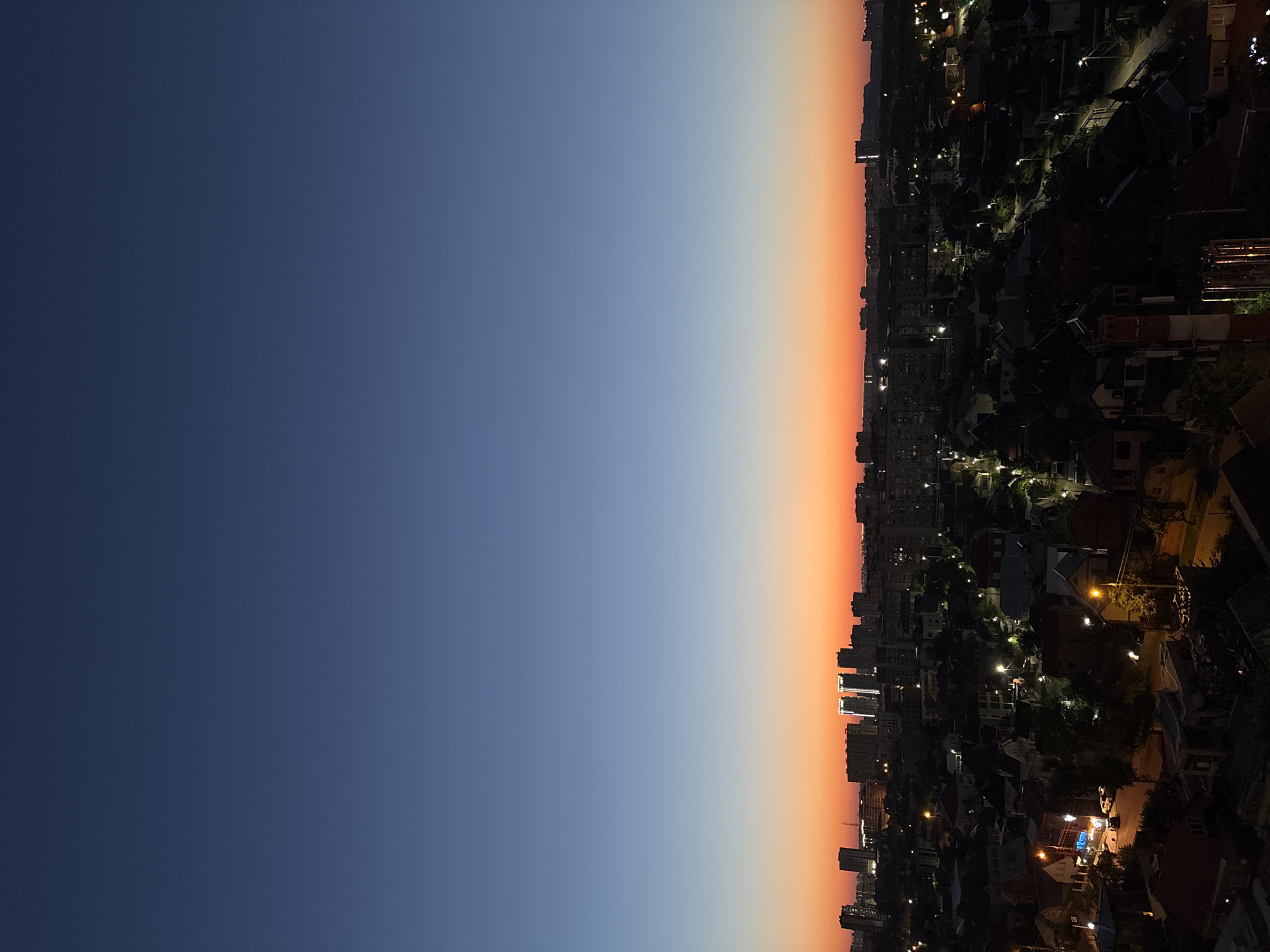 The image captures a cityscape at dusk or dawn, likely in Krasnodar, Russia, as indicated by the architectural style and urban planning. The sky transitions from a deep, clear blue at the zenith to a vibrant orange and yellow near the horizon, suggesting the sun is either rising or setting. Below the sky, a dense urban area is visible, characterized by a mix of mid-rise apartment buildings and smaller residential houses. Streetlights and illuminated windows indicate that the city is transitioning into night or waking up to a new day. The foreground features a collection of houses with dark rooftops and some visible vegetation, hinting at a residential neighborhood. The overall scene depicts a quiet urban environment during twilight hours, with the natural light of the sky contrasting with the artificial lights of the city. There are no discernible individuals or active events depicted in the frame. The perspective appears to be from a high vantage point, overlooking the city.