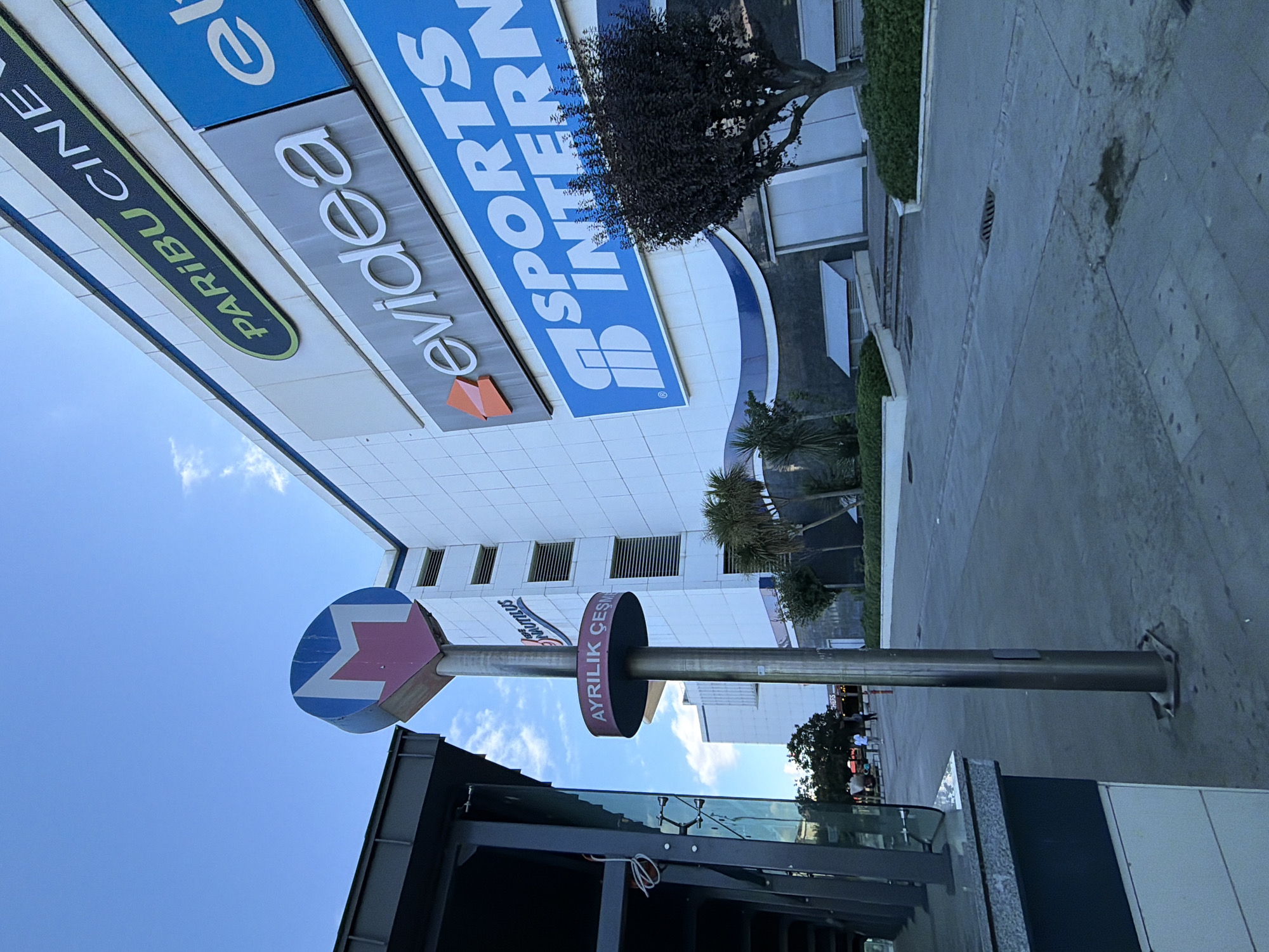 The image shows a street scene in Kadıköy, Turkey, with a metro station sign and a modern building in the background. The metro sign is circular with a stylized "M" and an arrow pointing downwards. Text on the sign reads "AYRILIK ÇEŞME". The building behind it is large and white, with several signs displaying company names and logos. One prominent sign reads "PARIBU CINEMA" and another "evidea". Below that is a large blue sign with white text that appears to say "SPORTS INTERN". The street in the foreground is paved, with a sidewalk and some greenery. A few people are visible walking in the distance, and a tree with dark foliage stands to the right of the frame. The sky is blue with a few scattered clouds. The overall impression is of a busy urban environment with modern infrastructure.