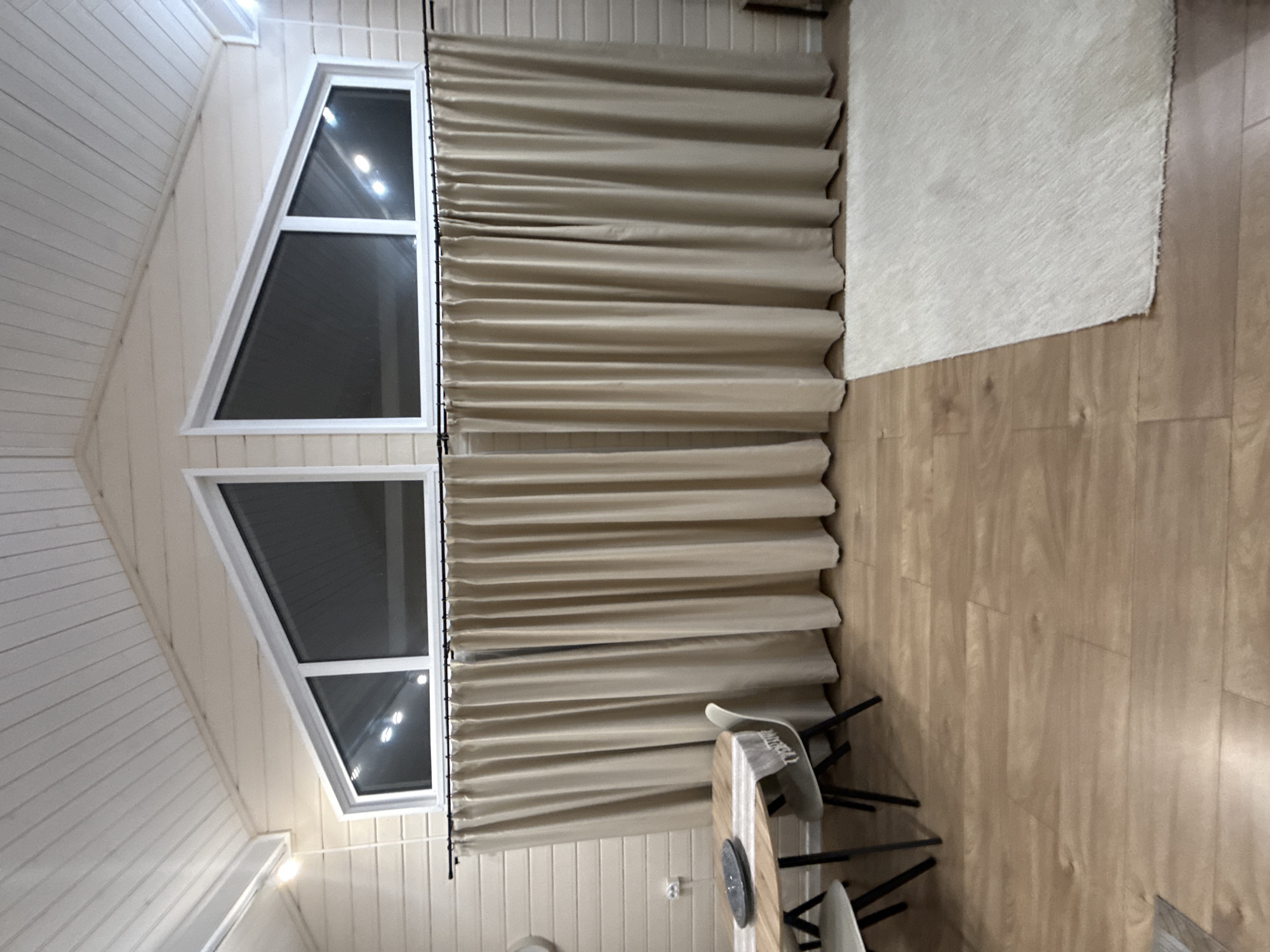 The image presents an interior view, likely taken in a residential space. Prominently featured are a set of windows with an unconventional triangular shape, which are covered by beige curtains. The curtains are gathered in folds, obscuring most of the light from outside.
To the right of the windows is a light-colored rug on a light brown hardwood floor.
Below the curtains, we see the corner of what appears to be a light-colored table with chairs around it, suggesting a dining area. The wall behind the curtains is beige vertical paneling. The overall impression is that of a room designed with a mix of modern and rustic elements, with a focus on light tones and natural materials. There is no verifiable information available to confirm the image location being in Buzhaninovo, Russia.
