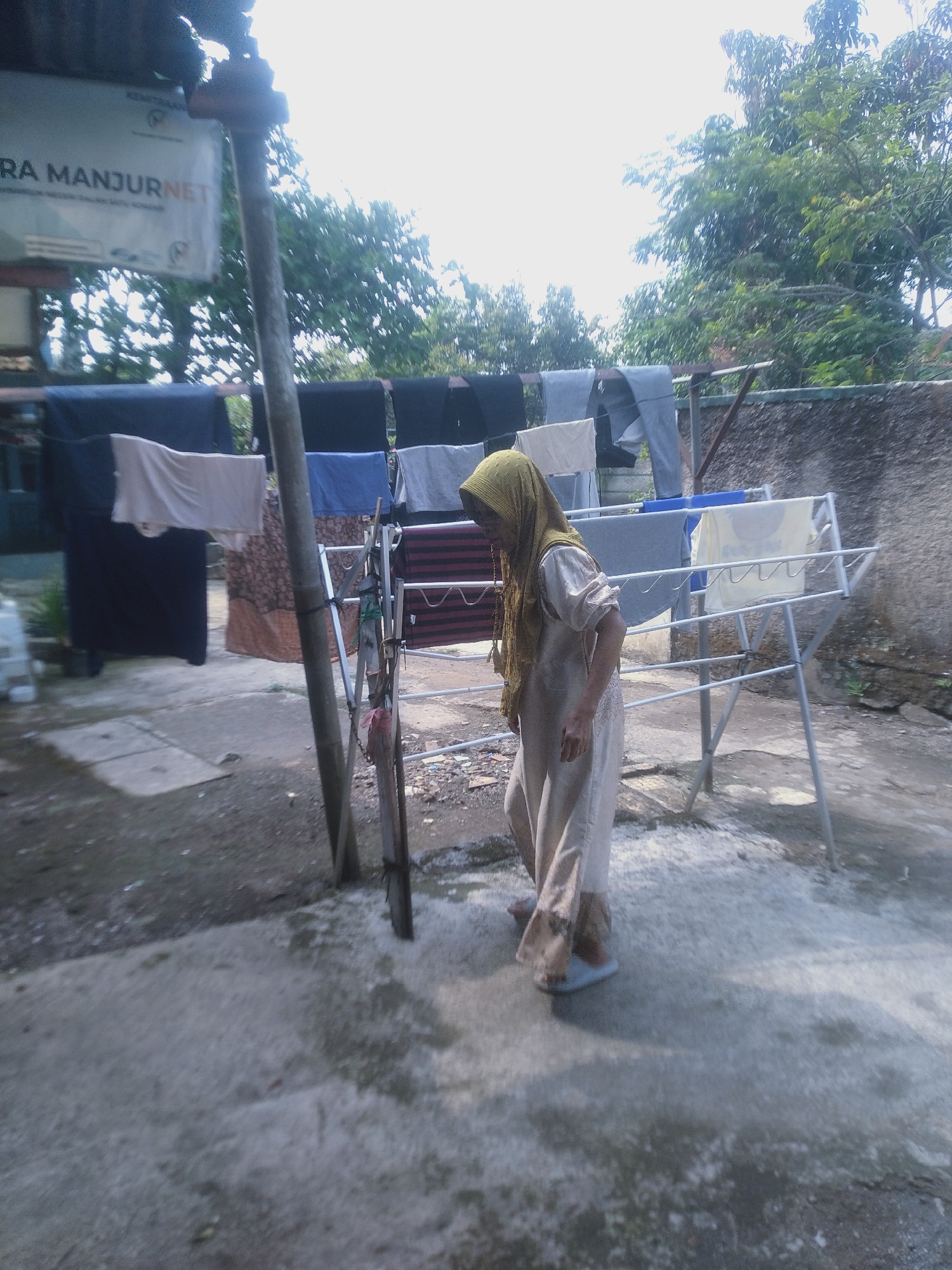 The image shows an outdoor scene in Bojong, Indonesia, likely a residential area. A woman, dressed in a light-colored headscarf and a long, flowing dress, is walking towards the right of the frame. She is wearing light-colored slippers. In the background, clothes are hanging on a drying rack, illuminated by bright sunlight. The drying rack is made of metal and has multiple tiers. Behind the rack, a textured concrete wall is visible, with trees and foliage beyond it. The ground in the foreground is a mixture of concrete and dirt, with some white specks scattered across it. A wooden pole is positioned in the center of the frame, supporting some of the hanging clothes. The overall atmosphere appears to be a typical day with laundry being air-dried.