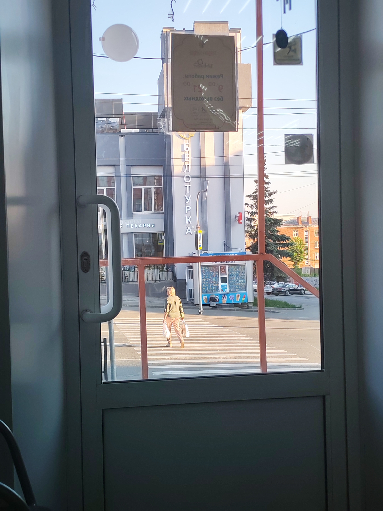 A view through a doorway shows a street scene in Izhevsk, Russia. A woman in a green jacket and light-colored pants walks across a crosswalk, carrying white plastic bags. Behind her is a small "MOROZHENOE" (ice cream) kiosk. Further back is a gray building with "ПЕКАРНЯ" (bakery) sign on it, and a white building with "БИБЛИОТУРКА" (bibliotourism) written vertically on the side. Cars are parked along the street, and buildings are visible in the background. The sky is clear. There are stickers attached to the glass doors.