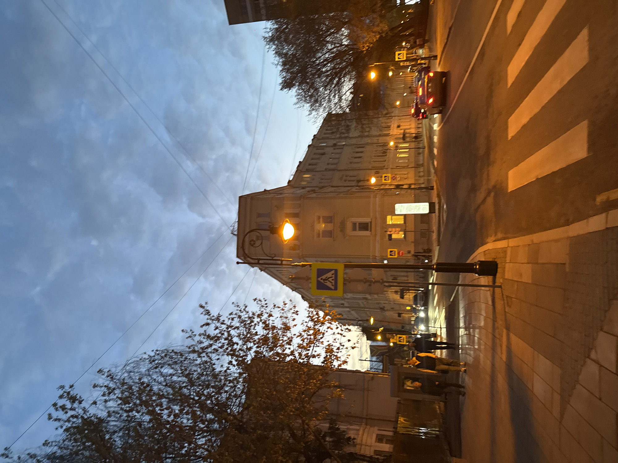 The image shows a street scene in Moscow, Russia at dusk. A tall, light-colored building dominates the left side of the frame, with street lights illuminating its facade. A pedestrian crossing sign is visible near the base of the building. A few people are walking along the sidewalk on the right side of the street. A car with its taillights on is driving on the road. The sky is cloudy. Bare trees are visible on either side of the street. A crosswalk is marked on the road.
