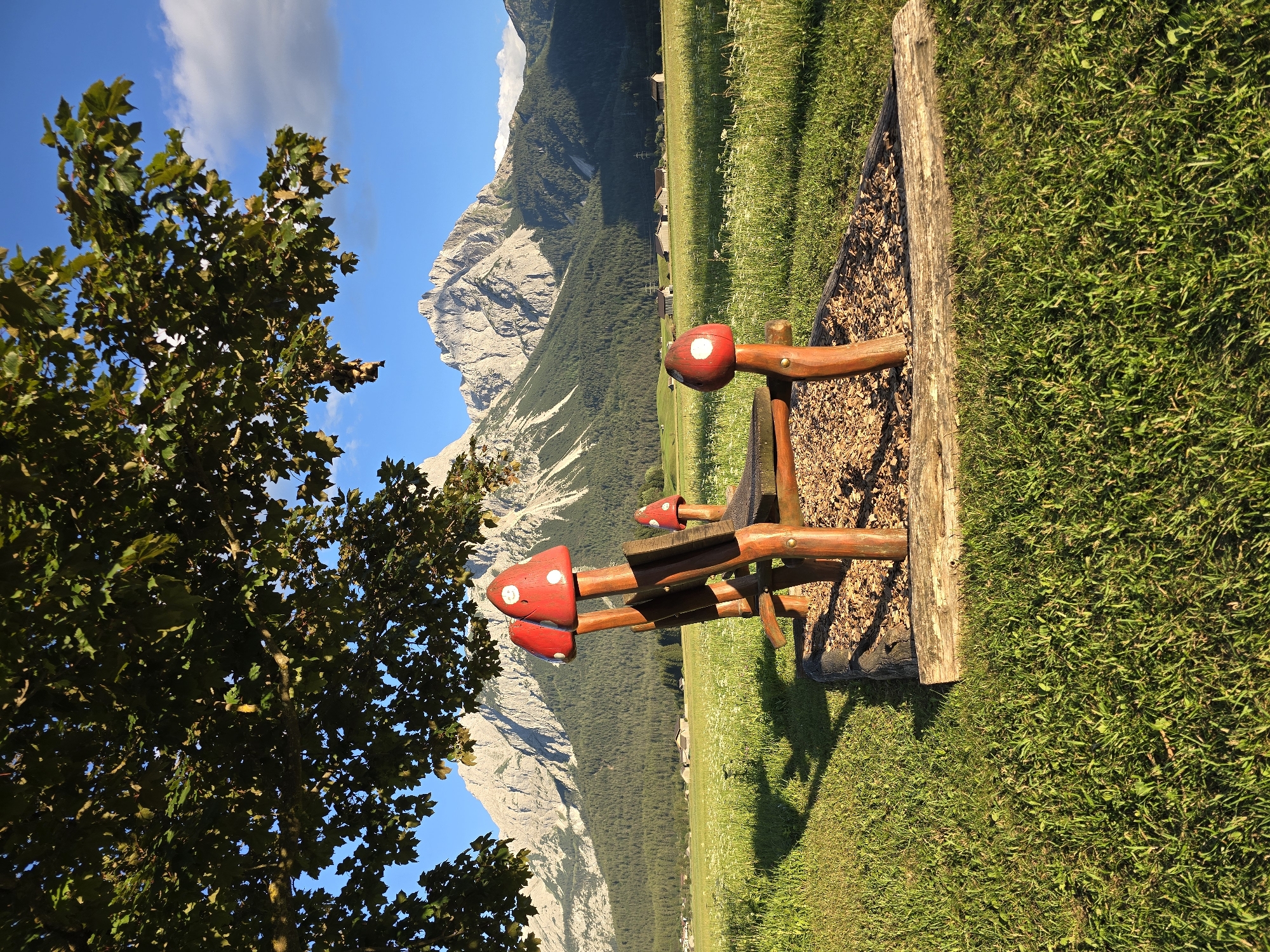 A whimsical wooden bench, adorned with bright red mushroom-shaped backrests and armrests, sits on a bed of wood chips in a grassy meadow. Behind the bench, a verdant forest stretches towards a majestic mountain range, its rugged peaks catching the golden light of the setting sun. The sky above is a brilliant blue, dotted with fluffy white clouds. In the distance, a scattering of charming farmhouses adds to the idyllic scenery. This picturesque setting is located in the charming Austrian village of Lermoos, nestled at the foot of the Zugspitze mountain.