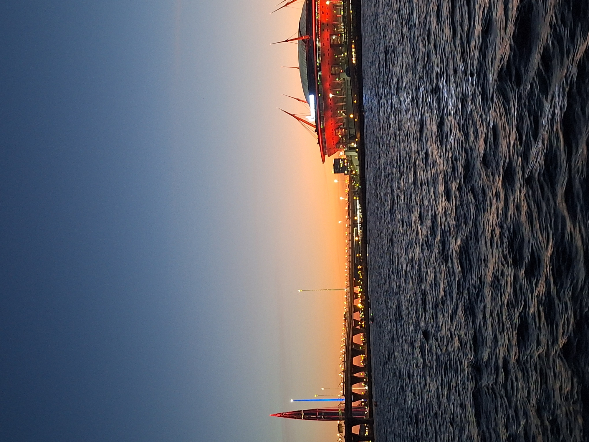 The image depicts a twilight view of the St. Petersburg skyline. In the foreground, dark, rippling water fills the bottom half of the frame, reflecting the faint glow of the sky and distant lights. Across the water, a wide bridge spans the scene, illuminated by numerous small lights along its railing. To the left of the bridge, a prominent skyscraper with a pointed red top and blue lights dominates the left side of the frame. To the right of the bridge, a large stadium with a distinctive red exterior and pointed roof structures is visible, also lit up. The sky transitions from a deep blue at the top to a warm orange and yellow gradient near the horizon, indicating either sunrise or sunset. The overall scene captures an urban landscape at dusk, featuring modern architecture and infrastructure over a body of water.