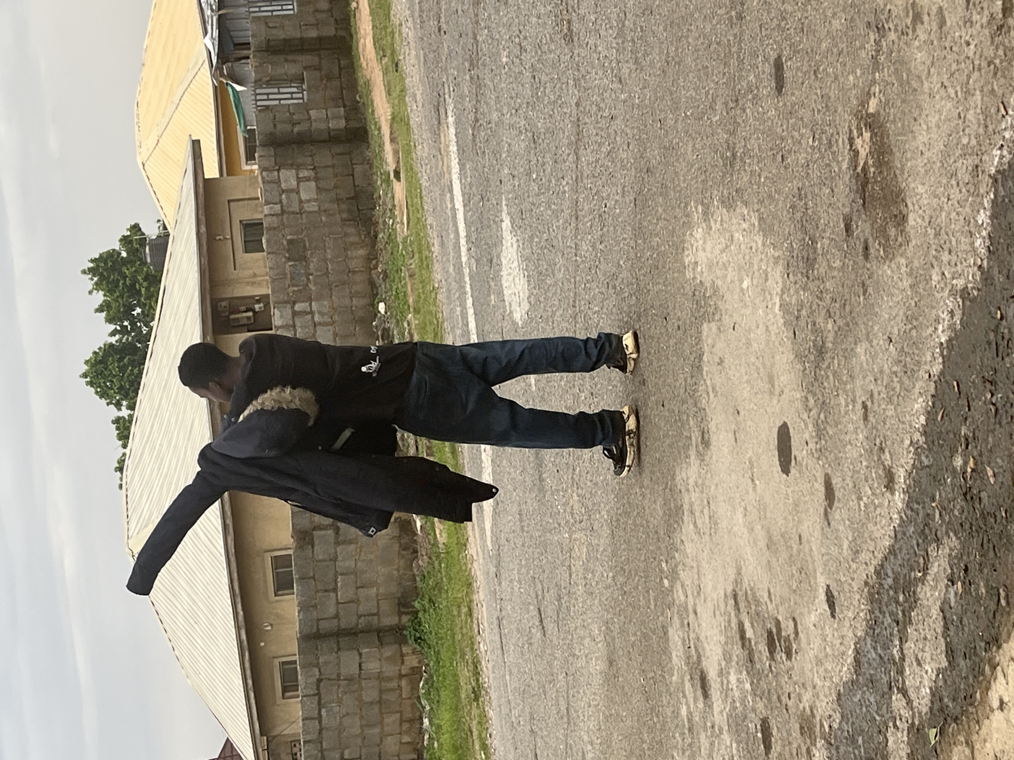 A man is standing in the middle of a road, facing away from the camera, with a dark jacket draped over his left arm. He is wearing dark blue jeans and black and white athletic shoes. The road is asphalt with some water patches and white markings. In the background, there is a low brick wall, followed by two houses with corrugated metal roofs, one yellow and one white. Trees are visible behind the houses. The scene appears to be in a residential area of Abuja, Nigeria. The sky is overcast.