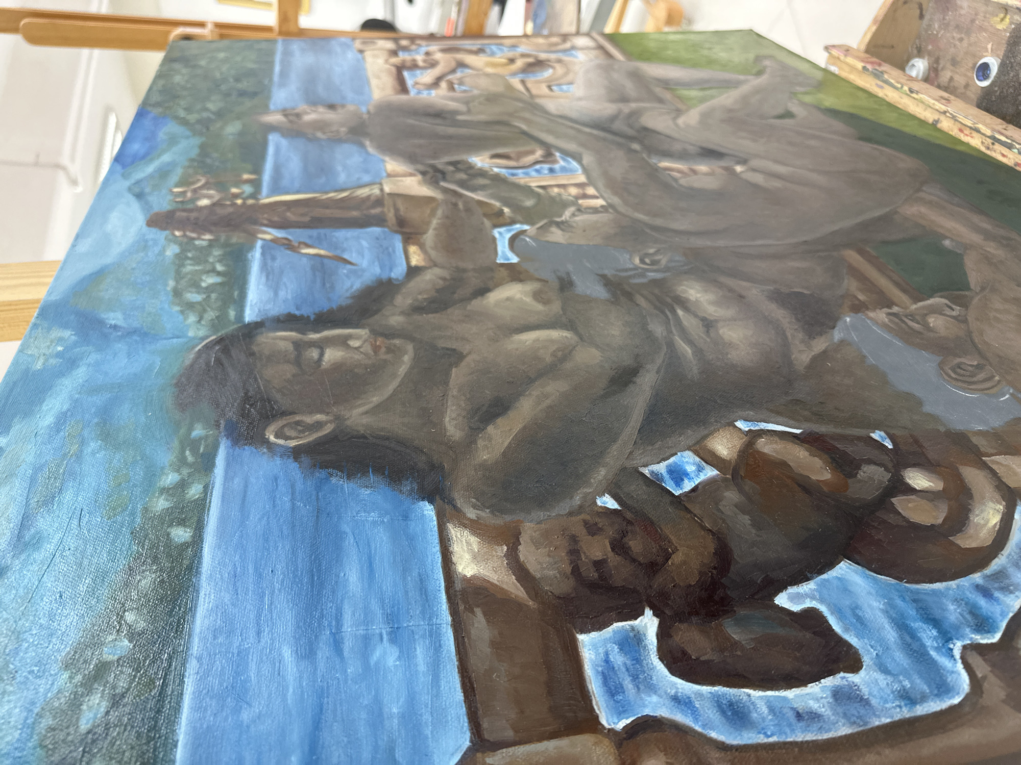 An angled, close-up photograph captures a large, rectangular canvas painting resting on a wooden easel within what appears to be an artist's studio in San Andrés Cholula, Mexico. The painting depicts an allegorical or mythical scene with multiple figures rendered in earthy tones against a backdrop of vibrant blues and greens.
In the foreground of the painting, a prominent figure, possibly a man, is shown sleeping or resting, with dark hair and a serene expression. Their body is partially draped, and they appear to be reclining on or near a brown, sculptural element. Behind this figure, another individual, seemingly larger or in a more elevated position, is depicted in motion, their form suggesting a sense of upward movement or descent. Other less defined figures or elements are visible further in the background.
The background of the painted scene features broad strokes of blue, resembling flowing water or abstract drapery, creating a waterfall-like effect vertically across the canvas. To the right of the painting's upper section, hints of green suggest foliage or a grassy landscape. The overall style of the painting utilizes strong chiaroscuro, giving the figures a sculptural and volumetric quality. The painting appears to be in progress, with some areas more developed and others showing preliminary washes of color. No text is visible in the image.