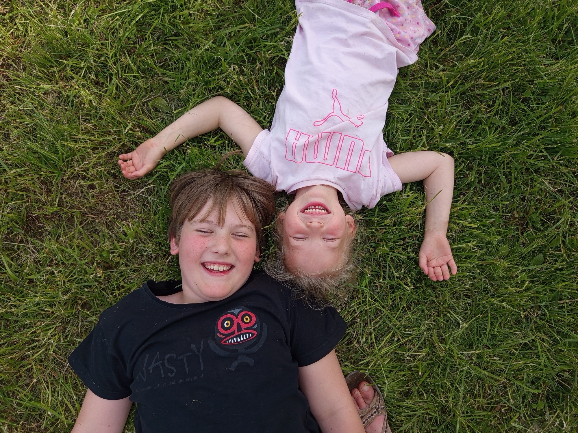 An overhead shot captures two children, a boy and a girl, lying on their backs in vibrant green grass in Ostrovtsy, Russia. Both children are smiling broadly with their eyes closed, conveying a sense of joy and amusement.
The boy, positioned in the lower left of the frame, has short brown hair and is wearing a black t-shirt. His t-shirt features the word "NASTY" in large white letters, beneath which smaller text reads "something that is ridiculously good." A graphic of a stylized red and yellow monster with prominent eyes and teeth is also visible on his shirt. He appears to be wearing open-toed sandals on his right foot.
The girl, situated in the upper right, has blonde hair and is wearing a light pink t-shirt with a pink Puma logo and the brand name "PUMA" printed on it. Her head is inverted relative to the boy, suggesting a playful pose. She is also wearing pink patterned pajamas or shorts.
The children are surrounded by lush, natural green grass, indicating an outdoor setting during daytime. The soft lighting suggests either an overcast sky or an early morning/late afternoon time. The overall scene depicts a carefree and happy moment shared between the two children.