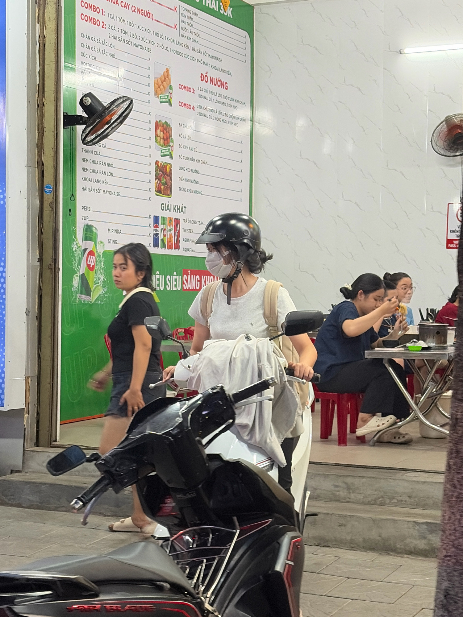 This is a street-level photograph taken at night in Da Nang, Vietnam. The scene depicts an outdoor dining area of a restaurant, with a menu board prominently displayed on the left. The menu lists various food and drink options, including "COMBO 1," "COMBO 2," "DO NUONG," and "GIAI KHAT." In the foreground, two motorbikes are partially visible, one in front of the other. A woman wearing a black t-shirt and shorts is standing to the left of the frame, looking towards the right. Another woman, wearing a white t-shirt and a helmet, is seated on a motorbike and looking forward. In the background, several people are seated at tables, eating and drinking. The environment appears to be a typical street in Da Nang, with concrete sidewalks and a busy atmosphere. The lighting suggests an urban setting at night, possibly from streetlights and the restaurant's interior lighting.