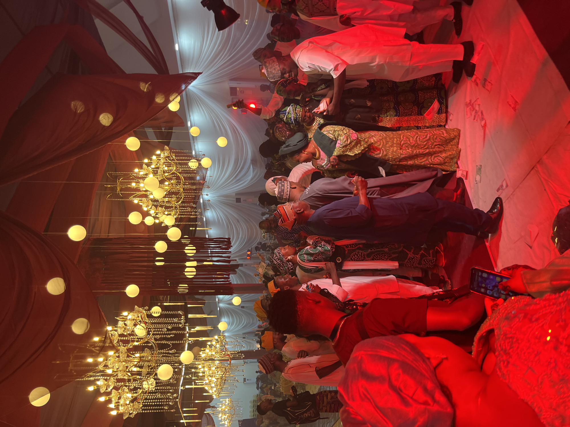 The image depicts a crowded indoor gathering with a festive atmosphere. The ceiling is adorned with large, ornate chandeliers and numerous spherical lanterns, draped with red fabric. The walls are covered in white fabric, creating a formal ambiance. In the foreground, a person, partially obscured by red lighting, is holding up a smartphone. The midground and background are filled with a diverse group of people, predominantly men, dressed in traditional attire, including kufis and elaborate garments. Some individuals are engaged in conversation, while others are observing the scene. Several men are seen receiving or giving money, indicated by bills scattered on the floor. The lighting is dramatic, with a strong red cast over much of the scene, particularly in the foreground. The overall impression is that of a celebration or event, possibly a wedding or cultural gathering.