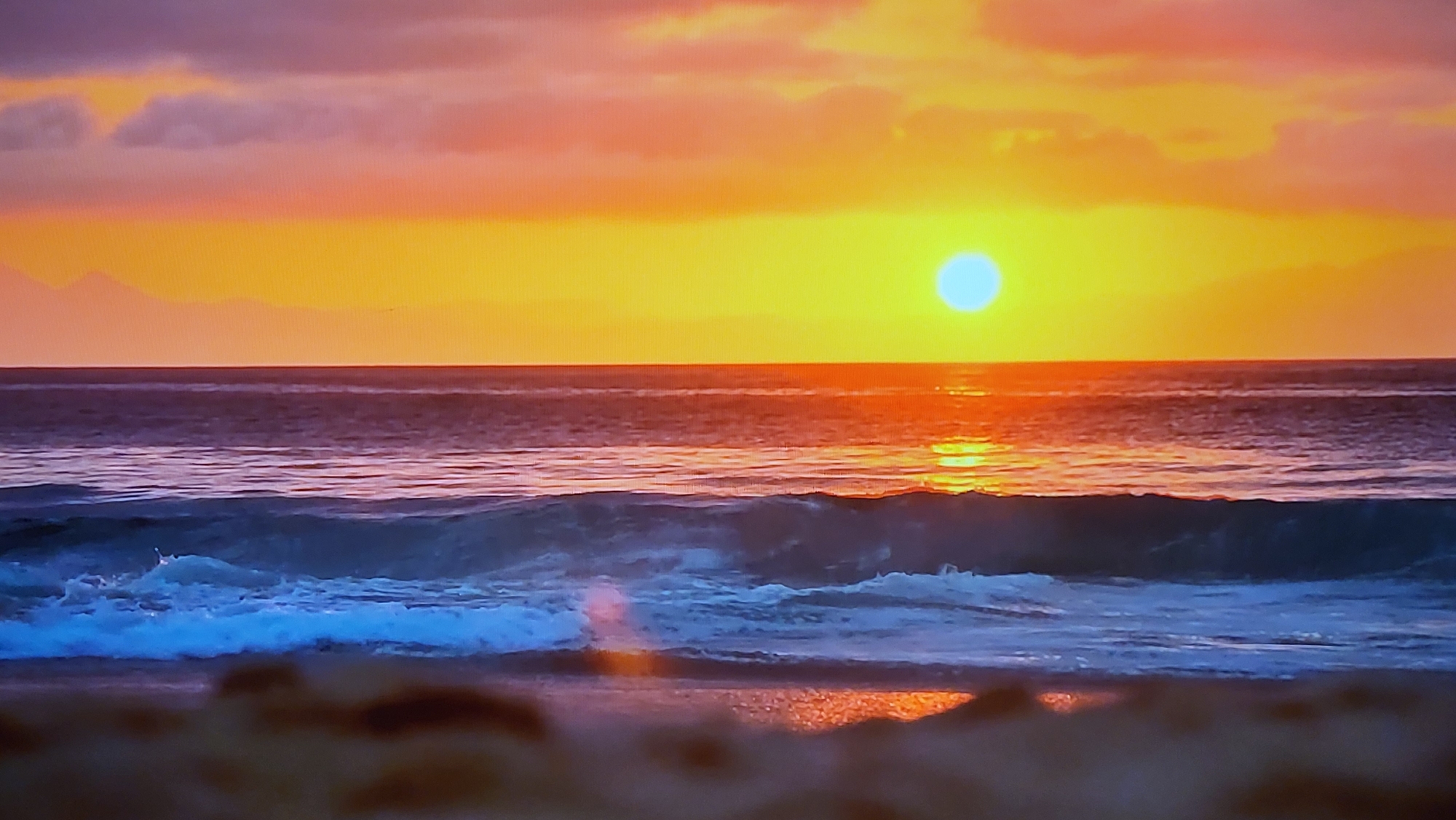 A vibrant sunset over a beach scene. The sky is filled with orange and yellow hues, with some clouds visible. The sun is a bright orb near the horizon, reflecting light onto the water. Waves are crashing onto the shore, with white foam visible. There is a dark mass visible in the far distance on the upper left. The foreground consists of the sandy beach.
This image does not depict Yerevan, Armenia, because Yerevan is landlocked. Therefore, no location in Yerevan, Armenia is applicable.