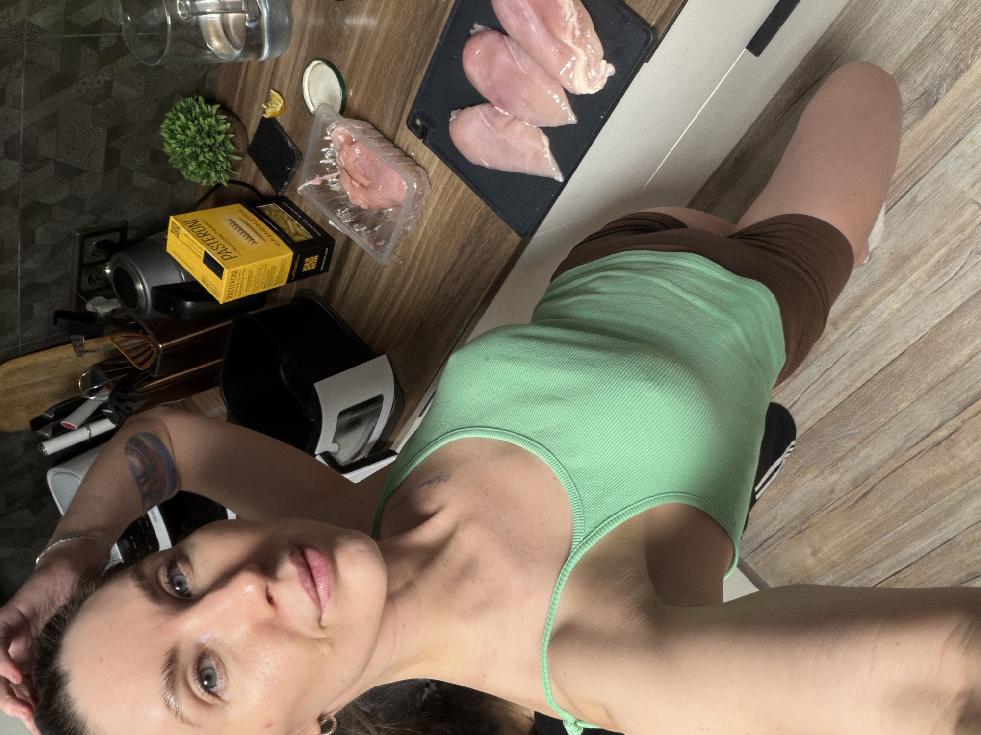 A woman is taking a selfie in a kitchen. She is wearing a green tank top and brown shorts. On the counter are raw chicken breasts, a package of sliced ham, a box of pasta, a glass of water, and a small potted plant. The kitchen appears to be in a residential setting. There is no indication of the city of Perm, Russia.