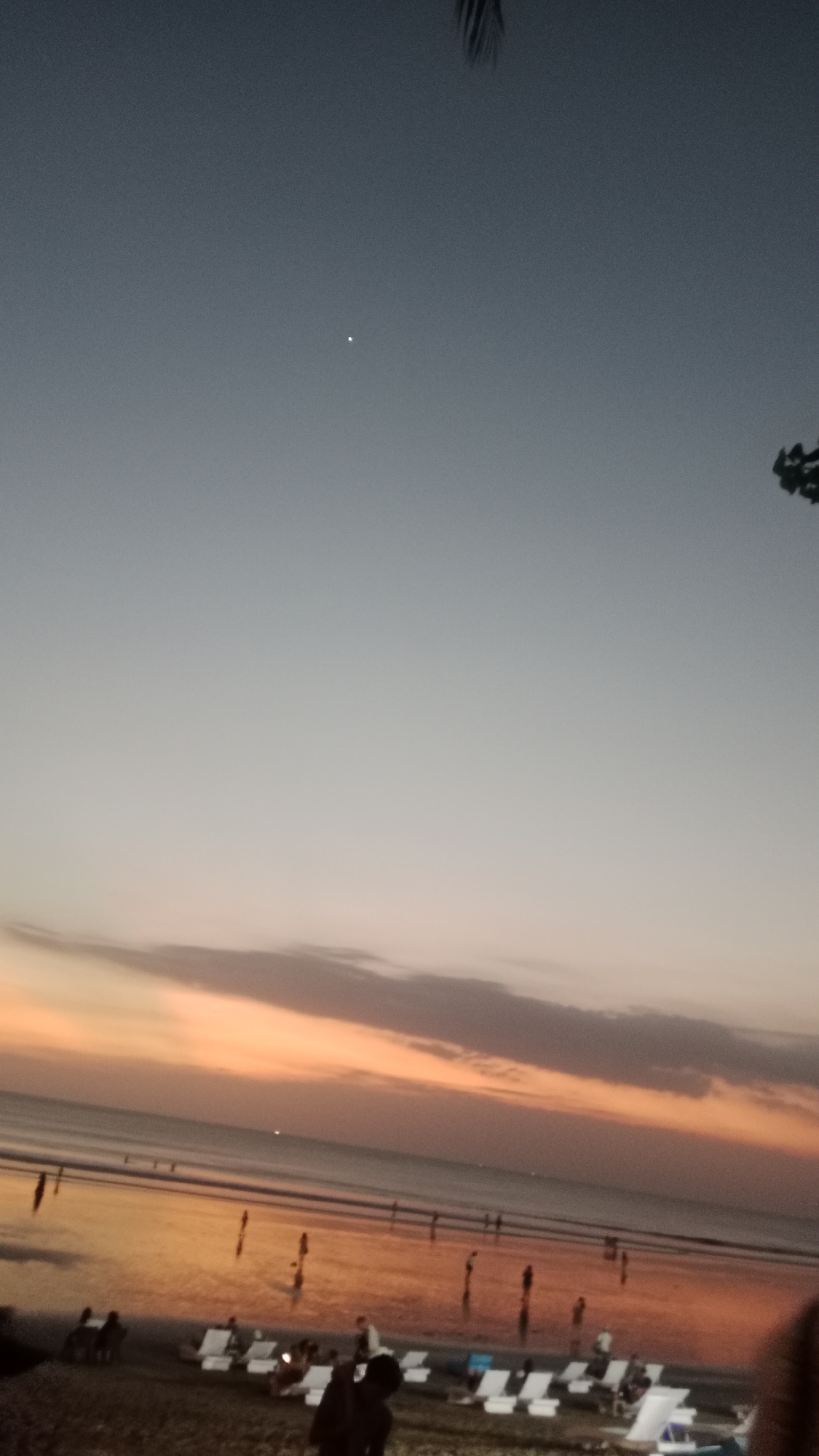 The image depicts a beach scene in Kuta, Indonesia, during twilight. The sky is a gradient of deep blue transitioning to a soft orange and pink near the horizon, indicative of sunset or sunrise. A faint star or planet is visible in the upper portion of the sky. The ocean is calm, with gentle waves lapping the shore.
On the wet sand of the beach, several figures are scattered, some appearing to walk or stand near the water's edge. Further back, a row of white lounge chairs are set up on the drier sand, some occupied by people. In the foreground, the silhouettes of people are visible, and parts of their bodies suggest they are casually observing the scene. The overall atmosphere is peaceful and serene. The location is Kuta, a well-known tourist destination in Indonesia, famous for its beaches and vibrant sunset views.