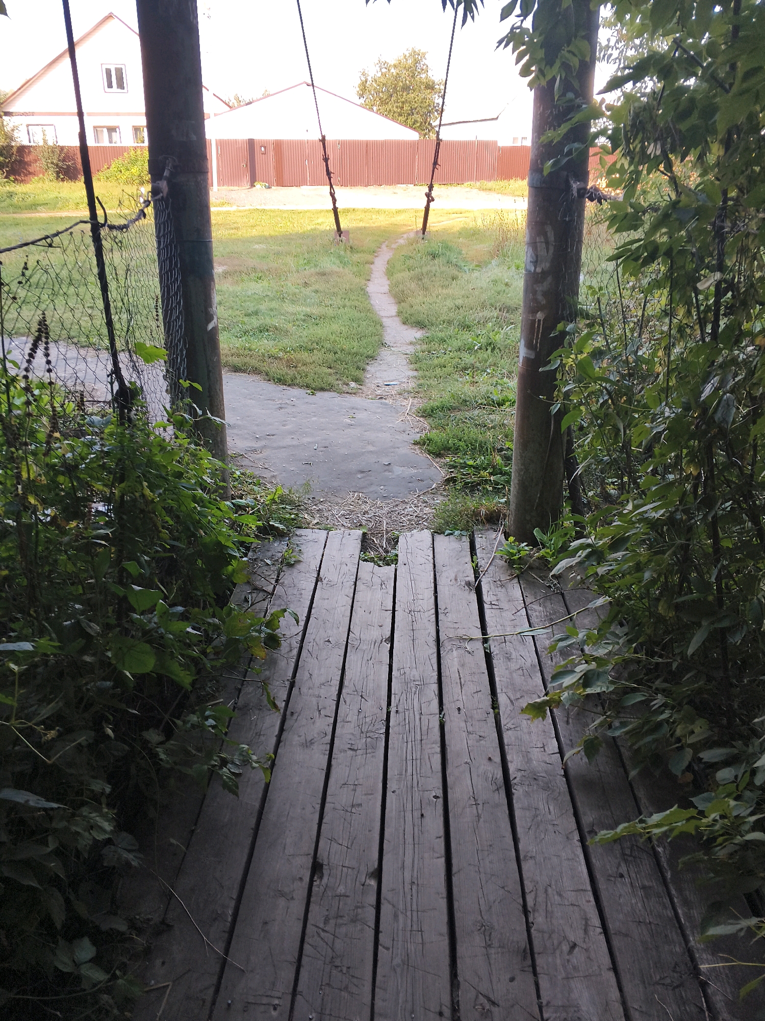 The image depicts an outdoor scene, likely a pathway leading towards buildings in Mokshan, Russia. In the foreground, a weathered wooden walkway composed of several planks runs diagonally from the bottom right to the bottom left. The planks are gray and show signs of age and use, with visible grain and some scratches. To the left of the walkway, a chain-link fence runs vertically, partially obscured by lush green foliage. Two large, rough-textured wooden posts stand on either side of the walkway, further framing the view.
Beyond the walkway and posts, a narrow dirt or gravel path curves through a grassy area. In the background, a light-colored house with a prominent gabled roof and windows is visible on the left, behind a reddish-brown corrugated metal fence. To the right of the house, another building with a similar fence is present. The sky is bright and appears to be overcast or the sun is low, casting a soft light on the scene. The overall environment suggests a rural or suburban setting with elements of nature and human construction. There are no people or animals visible in the image.