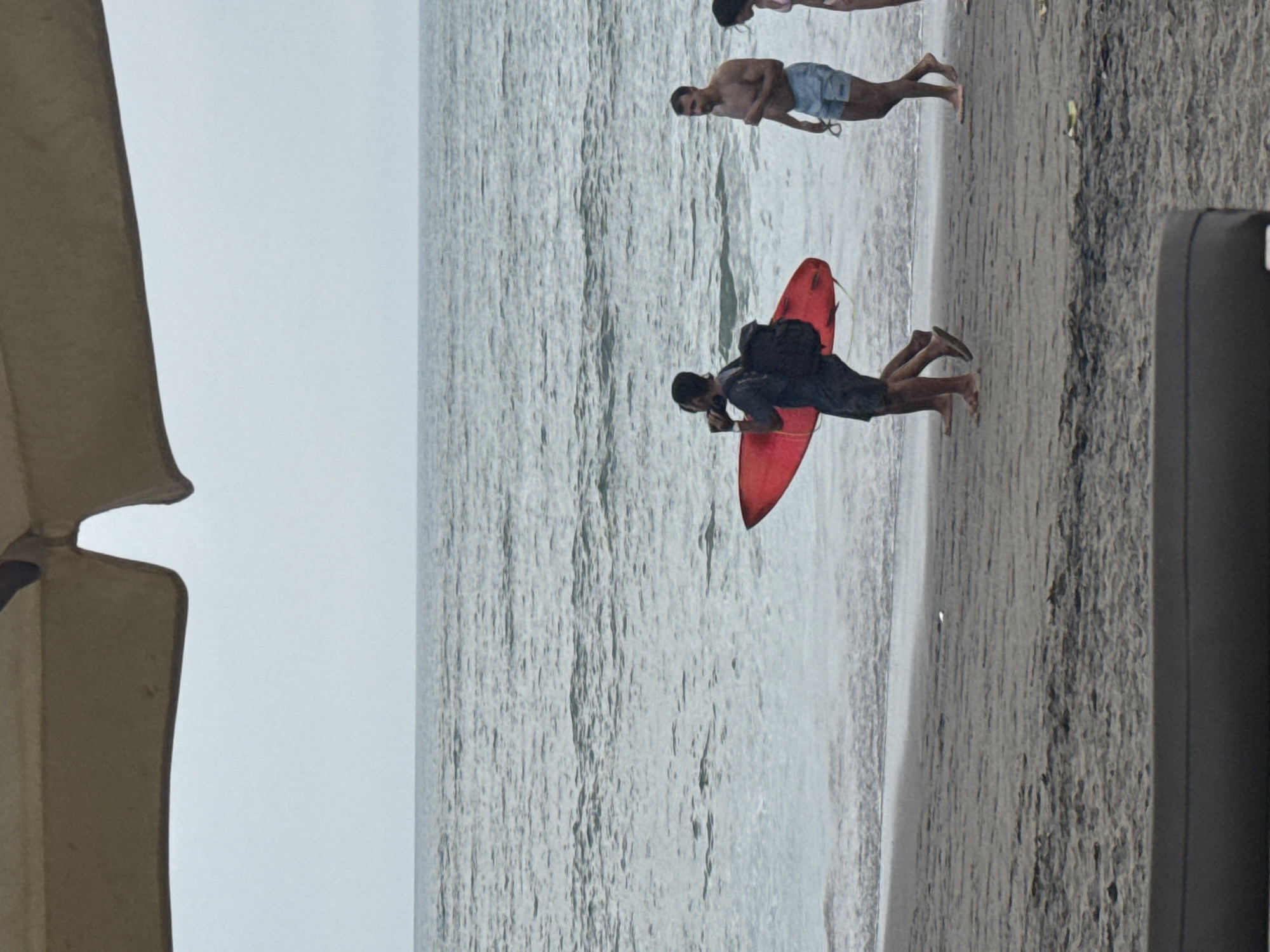 The image captures a beach scene in North Kuta, Indonesia, on an overcast day. The view is from the shore, looking out towards the ocean.
In the foreground, slightly to the left, a man is seen standing in the shallow water. He wears dark shorts and a dark top, with a black backpack slung over his shoulders. He holds a bright red boogie board (or bodyboard) with a visible leash, positioned upright beside him. His bare feet are partially submerged in the water.
Further out in the water, another man is standing, facing slightly towards the camera but looking out to sea. He is shirtless and wears light-colored swim shorts, possibly light blue. He appears to be holding a pair of goggles or sunglasses in his left hand. In the background, to the upper right, a third person is partially visible, only their lower body in light-colored swimwear is seen standing in the water.
The water is a muted grey-blue, with gentle waves breaking near the shore, suggesting calm conditions. The sandy beach, visible on the right, is a light tan color, showing subtle ripples from the receding tide. The sky is uniformly light grey, indicating an overcast or cloudy day, with soft, diffused lighting.
On the far left side of the frame, a large, light-colored fabric structure, likely part of a beach umbrella or canopy, is partially visible. On the far right, a dark, rectangular object, possibly a part of a beach chair or another piece of furniture, is also partially cropped. No specific emotions are discernible on the individuals, who appear to be enjoying the beach and water activities. There is no visible text in the image.