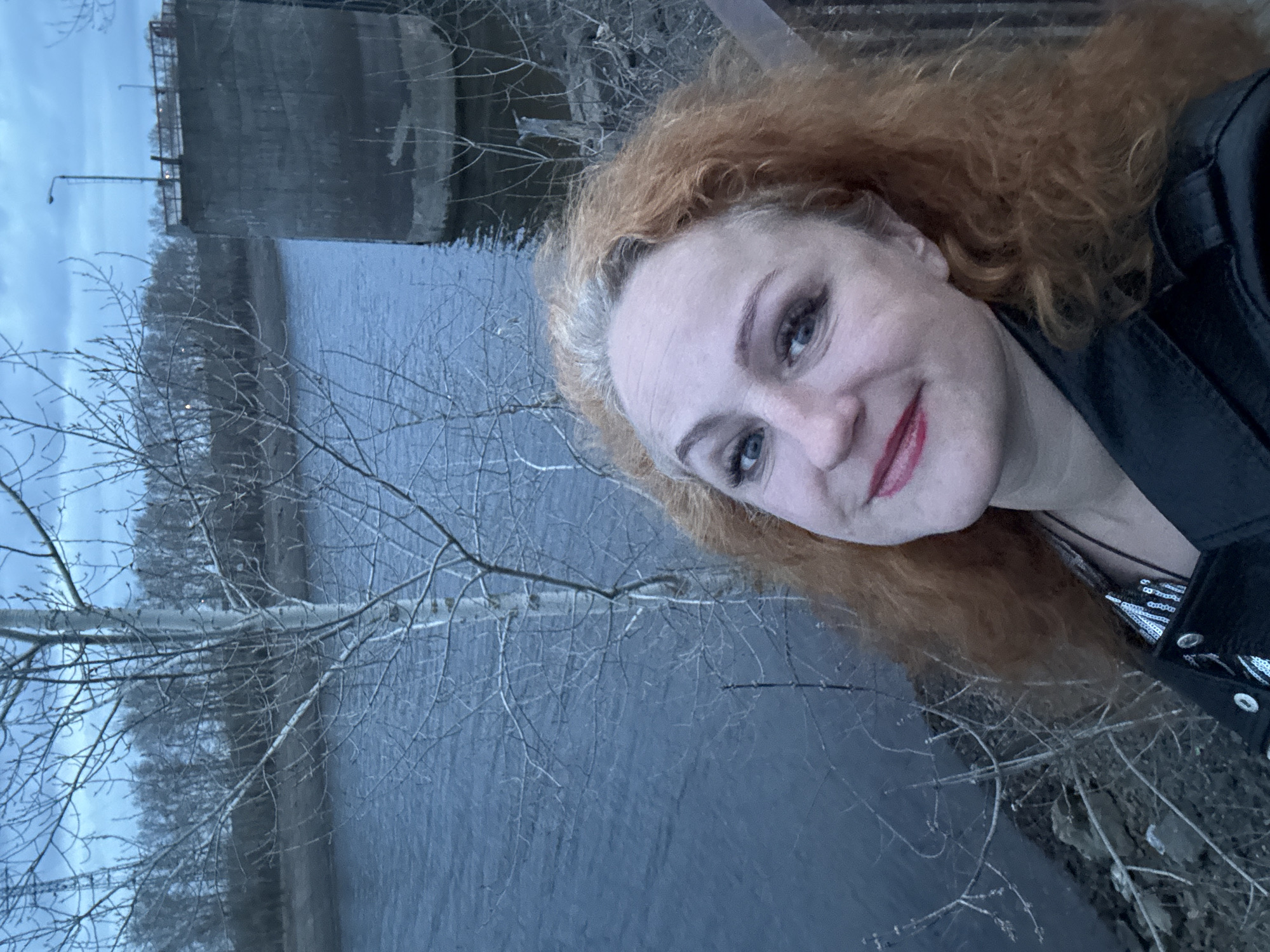 A woman with red, curly hair is taking a selfie. She is wearing a black jacket. In the background is a body of water and bare trees, suggesting a season like late fall or early spring. Further back, a concrete structure resembling a dam or bridge abutment is visible. Based on the location information (Chaikovsky, Russia), the water is likely the Kama River, and the concrete structure may be part of the Votkinsk Hydroelectric Station. The light suggests it is evening or early morning.
