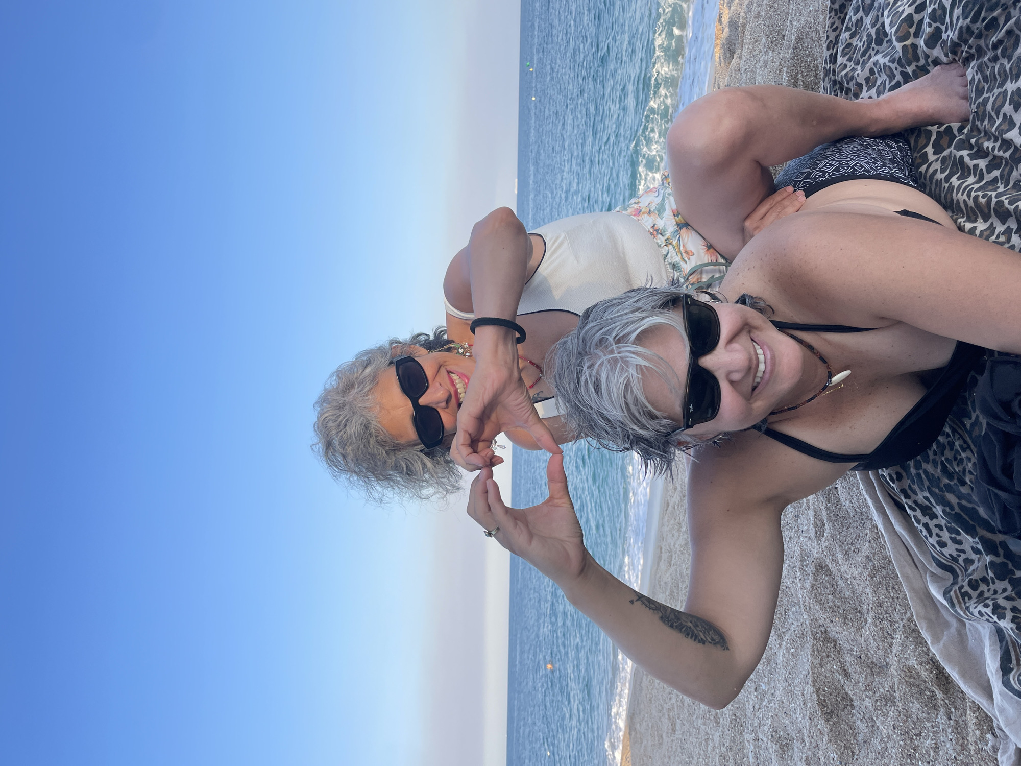 In Canet de Mar, Spain, two women with gray hair are on a beach. The woman in the foreground is lying on a patterned blanket on the sand, wearing a black bikini top and dark sunglasses. Her left arm is raised, and her hand is forming the bottom half of a heart shape. The woman behind her is kneeling on the sand, wearing a white tank top and floral shorts, also wearing dark sunglasses. Her hands are forming the top half of a heart shape, connecting with the other woman's hands. The ocean is visible in the background, with a clear blue sky above.