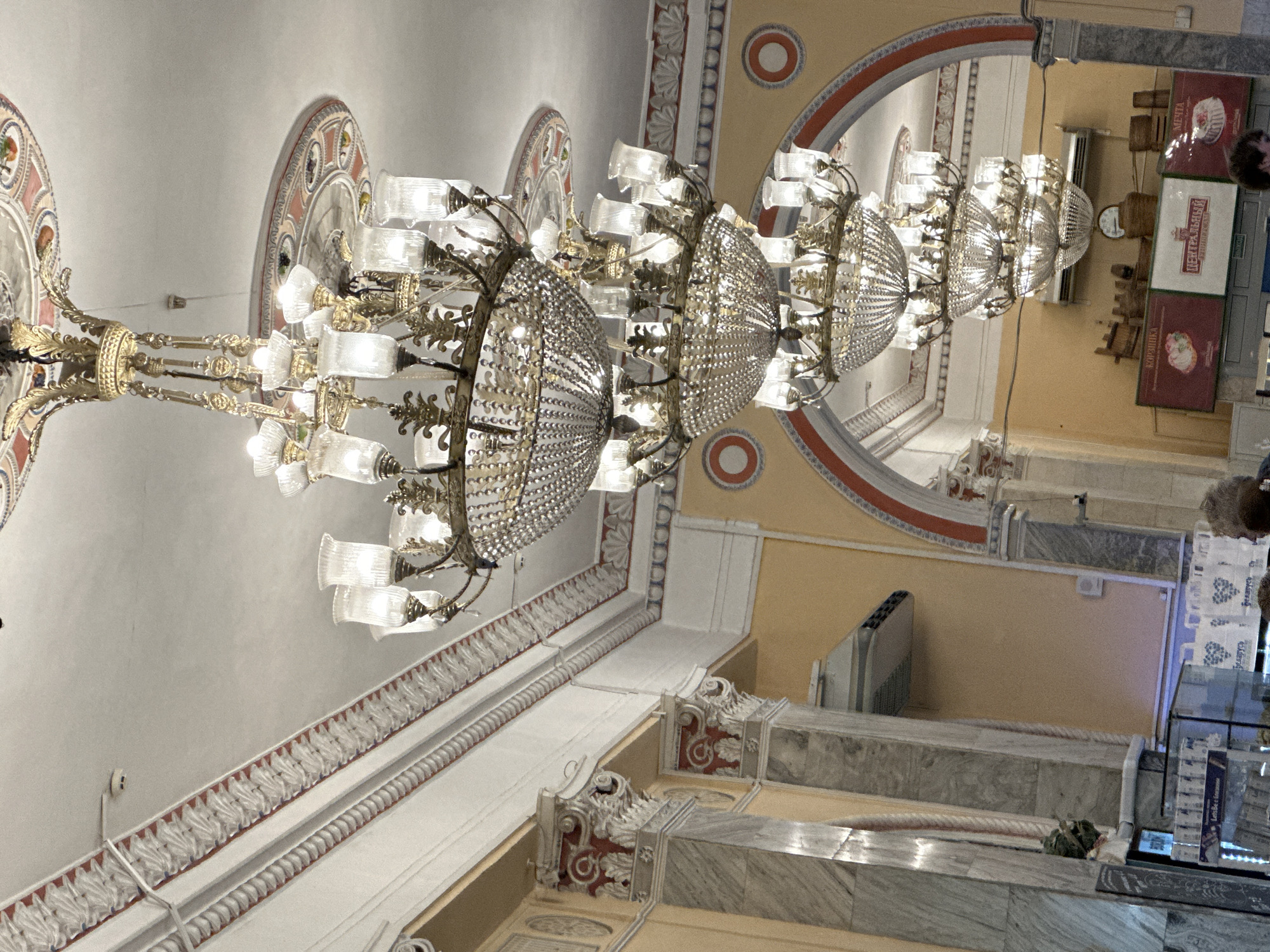 The image captures an interior view of what appears to be a large, ornate establishment, likely a cafe or restaurant, with a row of cascading crystal chandeliers hanging from a high, decorated ceiling. The architectural style features classical elements such as columns with red and white detailing and arched doorways. The walls are painted a warm yellow, adorned with decorative borders and circular motifs. In the foreground, to the left, a glass display case contains various items, and further to the right, white shopping bags with heart designs are visible. In the background, a clock and a heating/cooling unit are mounted on the wall.
This scene is located in the city of Minsk, Belarus, specifically within the Central Department Store (TSUM), a prominent shopping and cultural landmark. The chandeliers and decorative ceiling are characteristic of the store's historic and grand interior design.