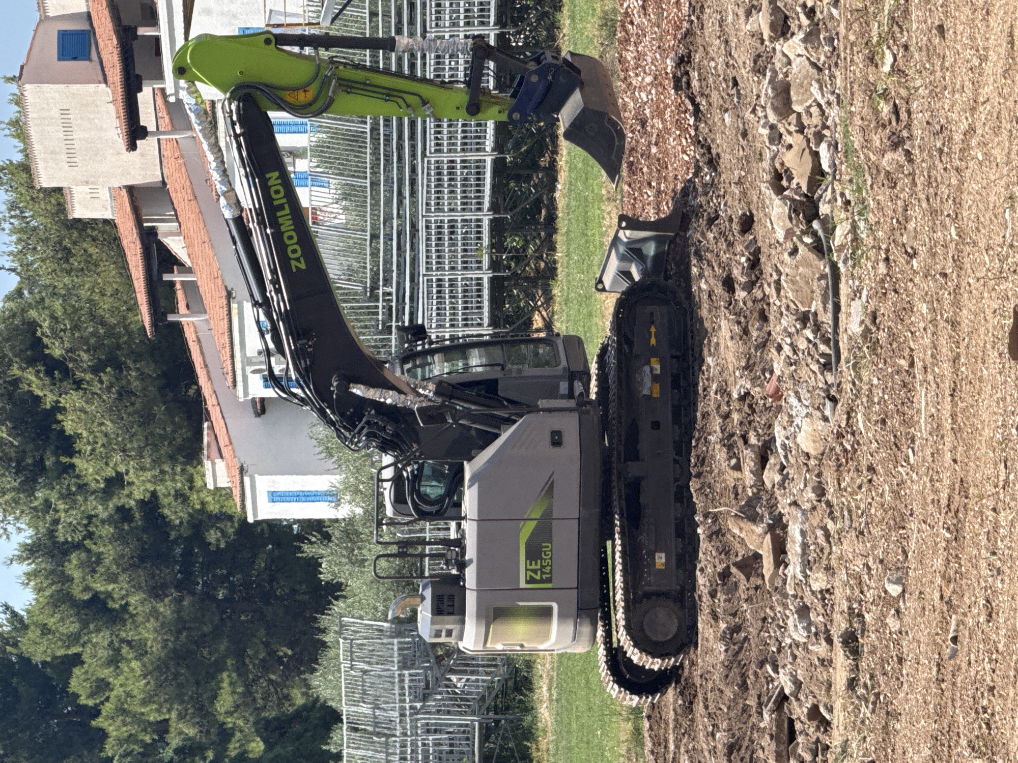 An excavator is parked on a dirt construction site, with a pile of dirt and debris in the foreground. The excavator is gray and green with the brand name "ZOOMLION" visible on the arm. In the background, there are tiered seating structures, green trees, and buildings with red tile roofs and white walls. The scene suggests ongoing construction work. The location is identified as Budva, Montenegro.