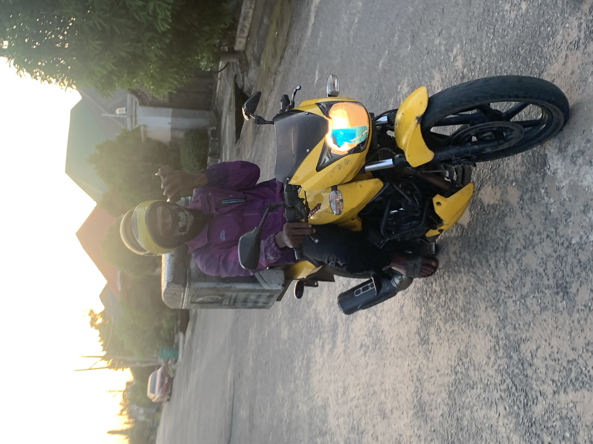 The image depicts an individual sitting on a yellow motorcycle with a black seat and tires on a paved road. The person is wearing a purple jacket and black pants. They also wear a yellow helmet, and one foot is covered by a black boot. The motorcycle has its headlight on. Behind the motorcycle and the person, there is greenery including trees. In the background, there are houses. The sun is visible in the distance, creating a bright glow in the sky. Based on the visible environmental features and the style of the motorcycle, this could plausibly be a street in Abuja, Nigeria.