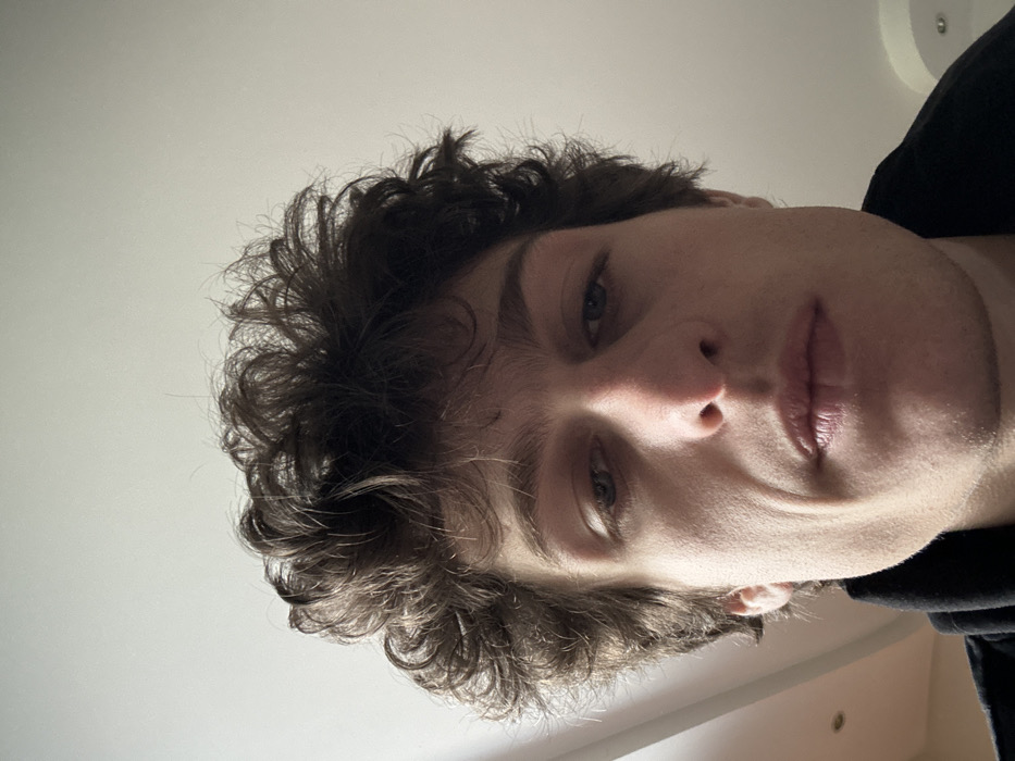 The image presents a close-up, low-angle portrait of a young adult male, likely in his twenties, with fair skin and dark brown, voluminous, curly hair. His gaze is directed slightly downward, and his expression is neutral. His mouth is closed, and his lips are full. Faint lines are visible on his cheeks near his nose and mouth. He is partially clad in a dark, possibly black, garment, the collar of which is visible at the bottom of the frame. The background consists of a plain, light-colored ceiling, with a subtle circular architectural detail or recessed light fixture visible towards the upper right. The scene is an indoor environment. The image itself provides no specific visual cues or landmarks to confirm the location within the city of Vincennes, France, as specified in the request.
