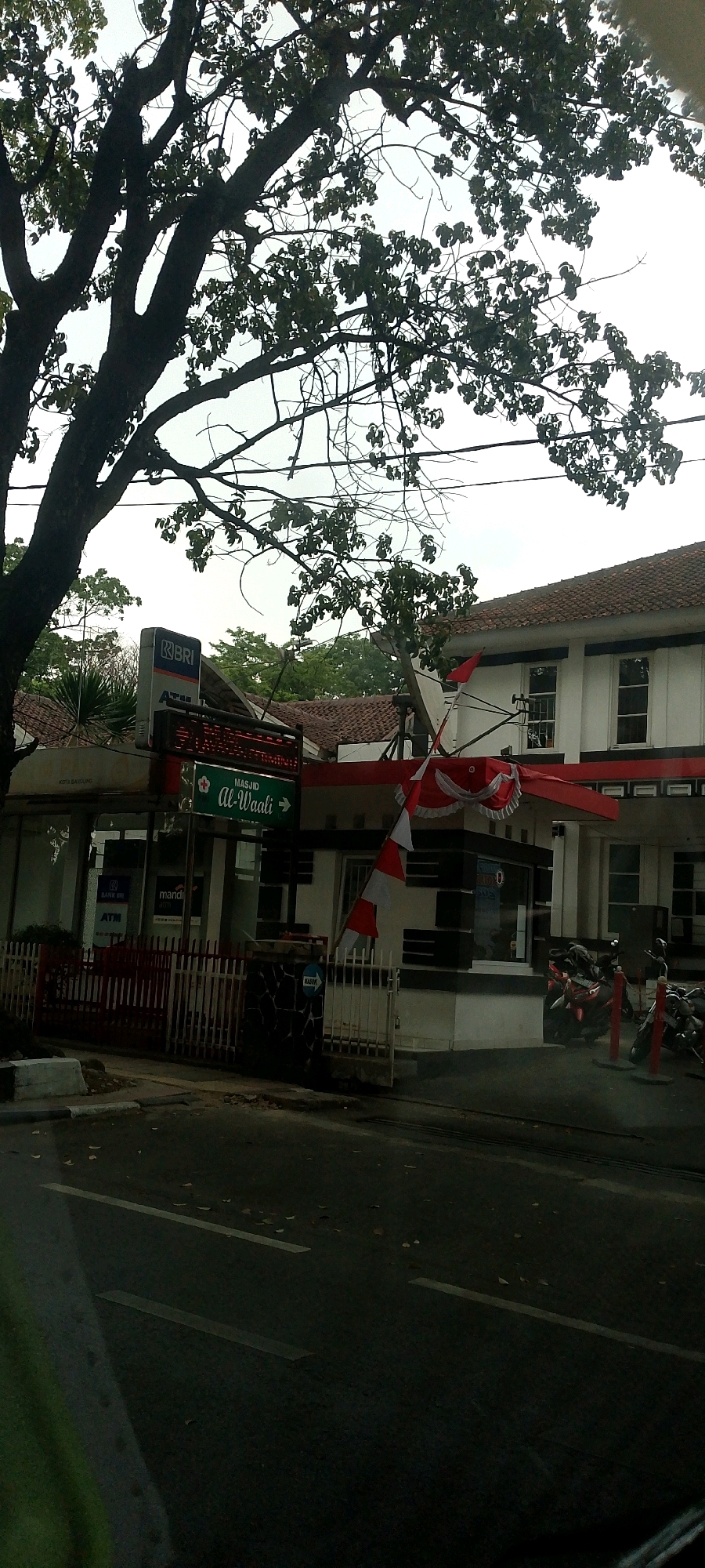 The image captures a street scene in Bandung City, Indonesia, with a focus on a building that houses a bank and a mosque. A large tree with dark branches and sparse leaves dominates the left side of the frame, obscuring the sky. The building, painted white with a red-tiled roof, has several windows and an awning adorned with red and white fabric decorations, possibly indicating a local celebration or national pride, given the red and white colors of the Indonesian flag.
In front of the building, a sign indicates "MASJID Al-Waali" with an arrow pointing to the right, suggesting the mosque is located in that direction. Below this sign, another sign displays "KOTA BANDUNG" and an emblem of the Bank Rakyat Indonesia (BRI) with an ATM. Next to the BRI ATM sign is a "MANDIRI" ATM sign. A red metal fence encloses the area in front of the building. Parked motorcycles are visible on the right side of the image, along the building's facade. The street in the foreground appears wet, reflecting some light, with scattered leaves on its surface. The overall lighting suggests it might be dusk or an overcast day.