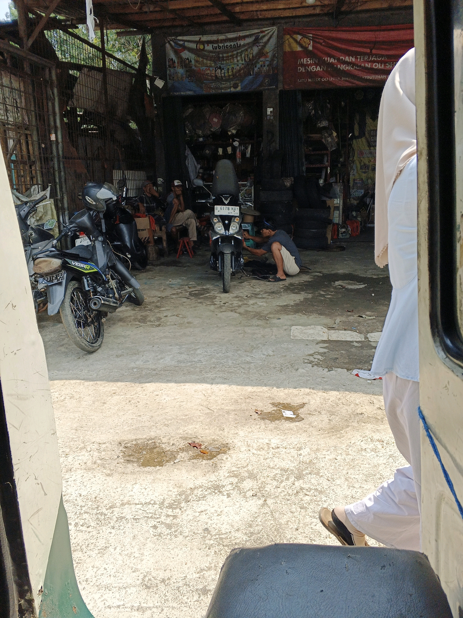 The image depicts a street-level view of a motorcycle repair shop in Warungkiara, Indonesia. The shop appears open-air, with a wooden roof supported by metal beams. In the foreground, a portion of a light-colored vehicle, possibly a passenger van or a trishaw, is visible on the left and right edges, indicating the photo was taken from inside.
Several motorcycles are present. On the left, a black motorcycle with a white and green accent and a license plate reading "F 2312 UU" is parked. Another black motorcycle is in the center, its license plate reading "B 6178 NXJ". A third black motorcycle is visible in the background.
Three men are inside the shop. One man, wearing a dark blue shirt and beige shorts, is crouched down and appears to be working on the central motorcycle. Two other men are seated in the background; one is wearing a checkered shirt and the other a cap and t-shirt. They are observing the activity or conversing.
The ground is concrete and appears dusty and somewhat wet in places, suggesting recent rain or a cleaning. Various tools and spare parts are visible throughout the shop, stacked on shelves or laid out on the ground. In the background, a large red banner with Indonesian text is hung, along with other advertising materials. The environment suggests a functional, working repair shop in a semi-urban or rural setting.
