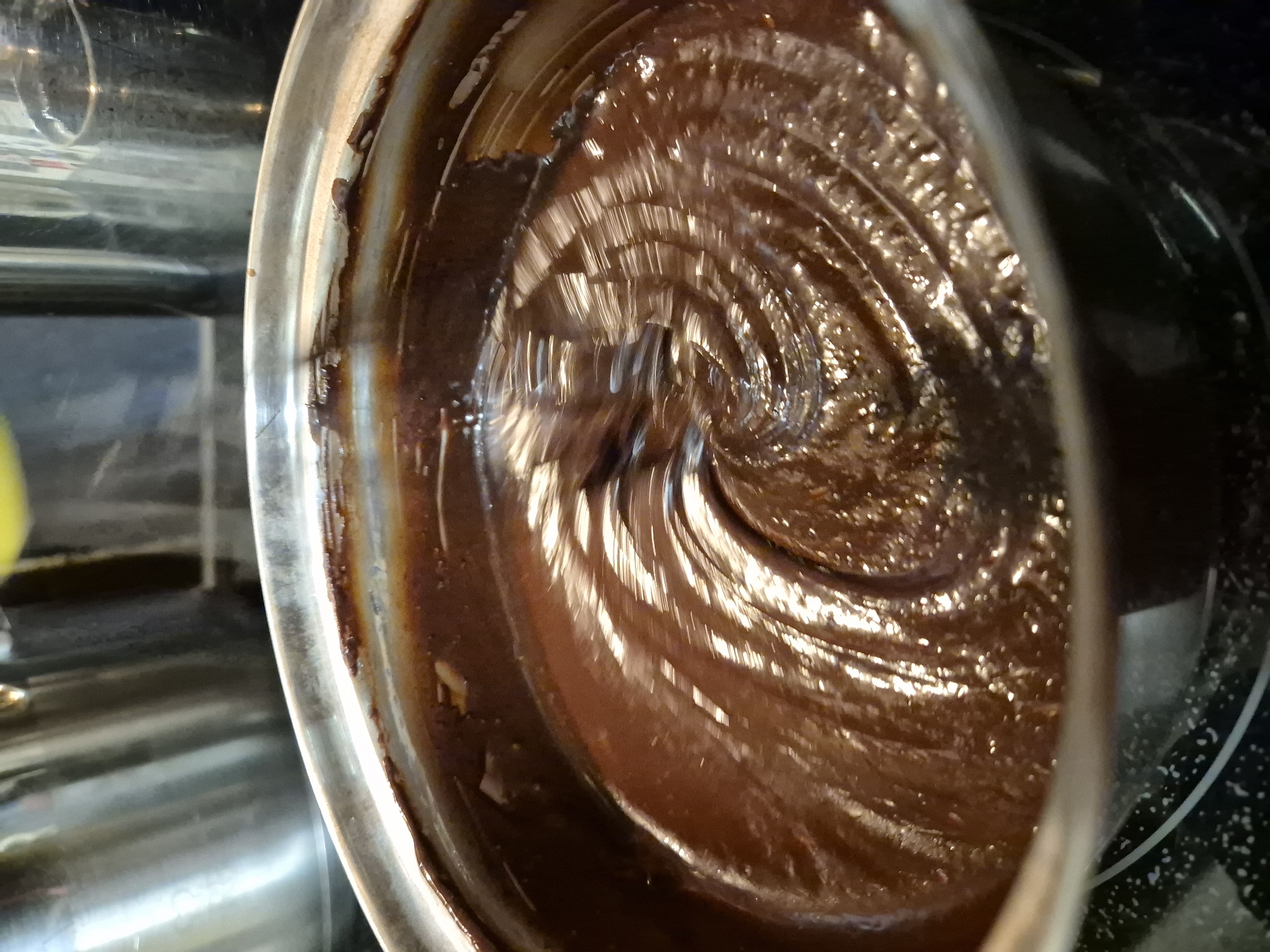 A close-up shot focuses on a silver mixing bowl containing a dark brown, glossy substance, likely chocolate batter or ganache. The mixture has been stirred, creating a swirling pattern. The bowl sits on what appears to be a black stovetop with white markings. To the left of the bowl, a stainless-steel surface is visible, reflecting the scene. The image suggests food preparation, specifically baking, but location within West Suffolk is not discernible.