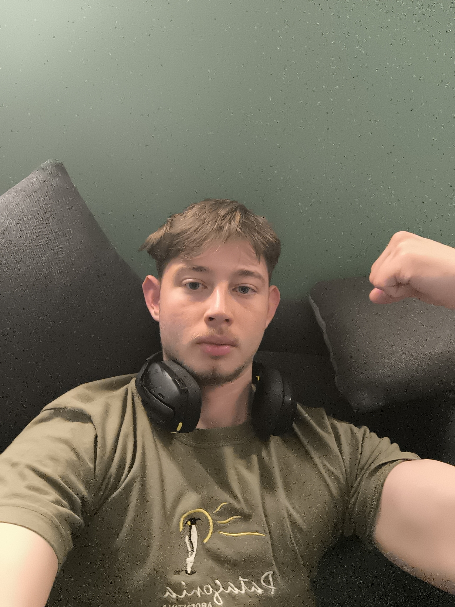A young man with light brown hair and light eyes is featured in a selfie taken indoors in Sofia, Bulgaria. He is wearing an olive green t-shirt with a graphic depicting a stylized figure under a yellow sun, and black over-ear headphones are around his neck. The man looks directly at the camera with a neutral expression, his right arm bent and fist clenched in a flexing pose, suggesting confidence or playfulness. He is seated on a dark grey couch, with parts of its cushions visible on either side of him. The background is a solid, muted greenish-grey wall. The lighting is soft and appears to be artificial, typical of an indoor setting. Reversed text on his shirt beneath the graphic partially reads "Patagonia".