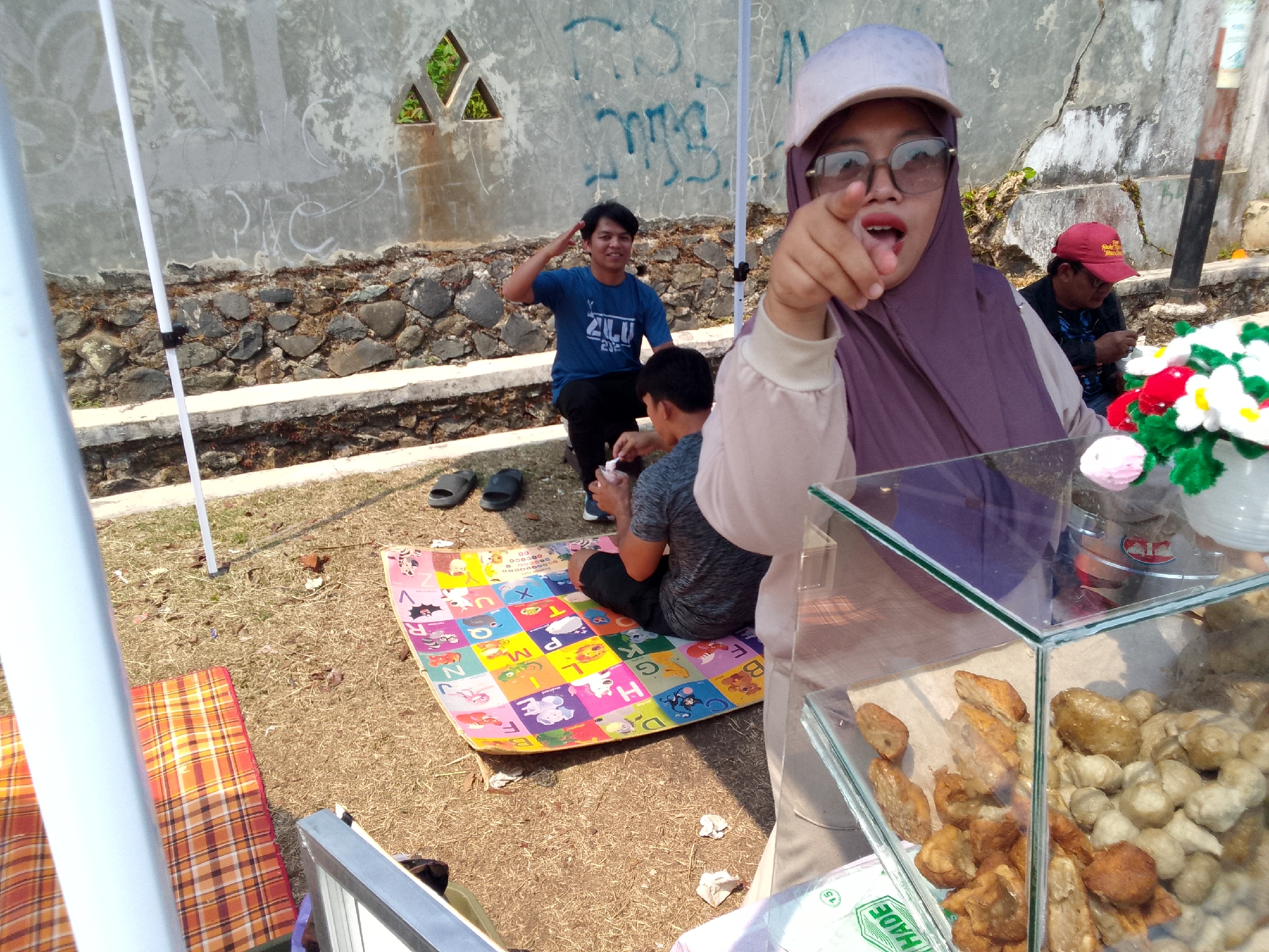 In Mandalawangi, Indonesia, a group of people are gathered outdoors. A woman in a purple hijab and a light-colored cap, wearing glasses, points towards the camera with an open mouth. She is in the foreground, to the right of the frame. Behind her, a man wearing a blue t-shirt with "ZULU" written on it salutes. Another person, partially visible, is seated on a colorful alphabet mat on the ground. There is a glass display case filled with fried food items, possibly snacks, in the foreground. The environment includes a rough stone wall, dry grass, and a white pole, suggesting an informal outdoor setting, possibly a market or a roadside stall.