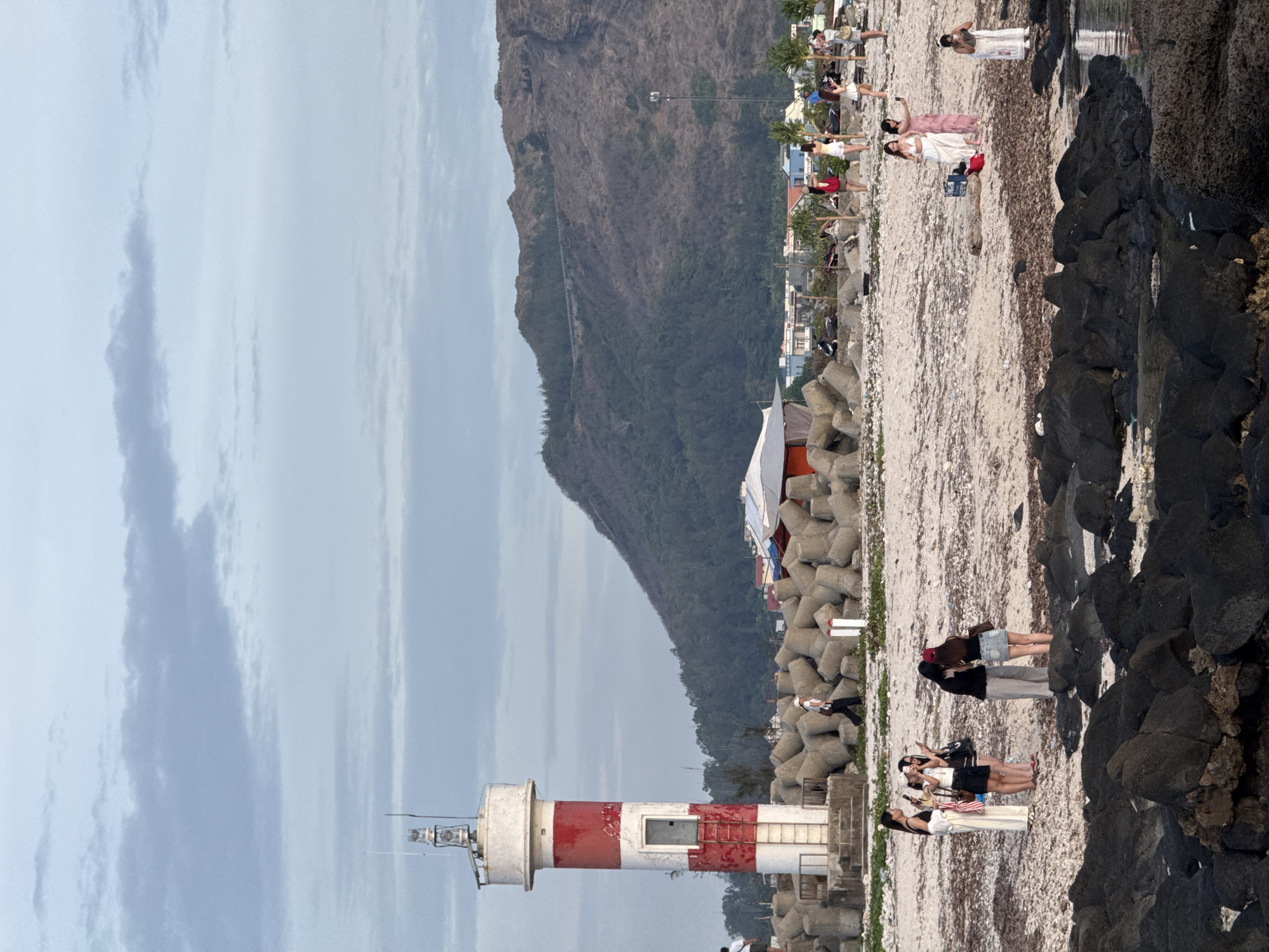 In the city of Lý Sơn Special Administrative Region, Vietnam, a scenic coastal view unfolds with a prominent lighthouse. The lighthouse, painted in alternating bands of red and white, stands tall on a concrete platform surrounded by large, interlocking concrete tetrapods, likely serving as a breakwater.
The beach is a mix of sand and small stones, with a few dark, jagged rocks visible in the foreground near the water's edge. A body of water, possibly the sea, is present to the right, reflecting the overcast sky.
Several individuals are scattered across the beach. On the left, a group of four young women are gathered, some taking photos with their phones. Further to the right, a group of women, some in light-colored dresses, are seen standing and interacting. One individual in a white dress is positioned near the water. In the middle ground, a solitary figure walks along the beach, carrying a bag. A few palm trees are sparsely planted on the sand.
In the background, a rugged, tree-covered mountain slopes upwards. A road is visible winding its way up the mountainside. A collection of buildings, appearing to be houses and some small structures with awnings, are situated at the base of the mountain, suggesting a small town or village. The sky is cloudy, with layers of gray and white clouds. The overall impression is a casual, late afternoon scene at a Vietnamese coastal location.