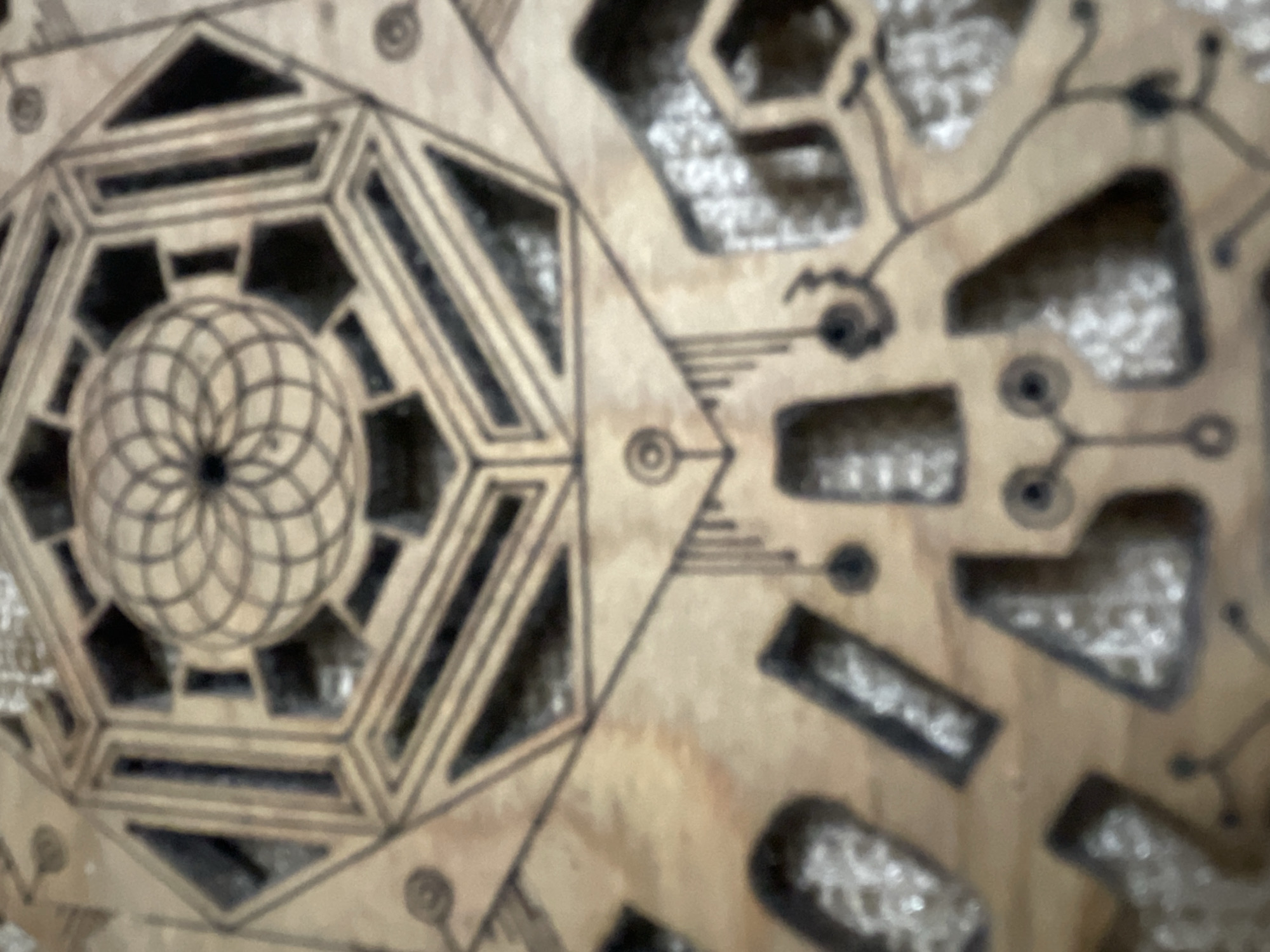 The image shows a close-up, blurry view of a detailed wooden carving. The carving features intricate geometric patterns, including a central spherical or geodesic dome-like design within a hexagonal frame. Below this, there are various abstract shapes and lines, some appearing as cut-out sections within the wood, filled with a white, possibly glittery material. The carving has an earthy, natural wood tone with dark lines outlining the designs. The overall impression is one of a complex, decorative object.
The provided context of Bhagsu, India, suggests this carving might be a piece of local art or craft, possibly found in a shop, home, or religious site within the town. However, without further information or visual cues, it is impossible to pinpoint its exact location within Bhagsu.