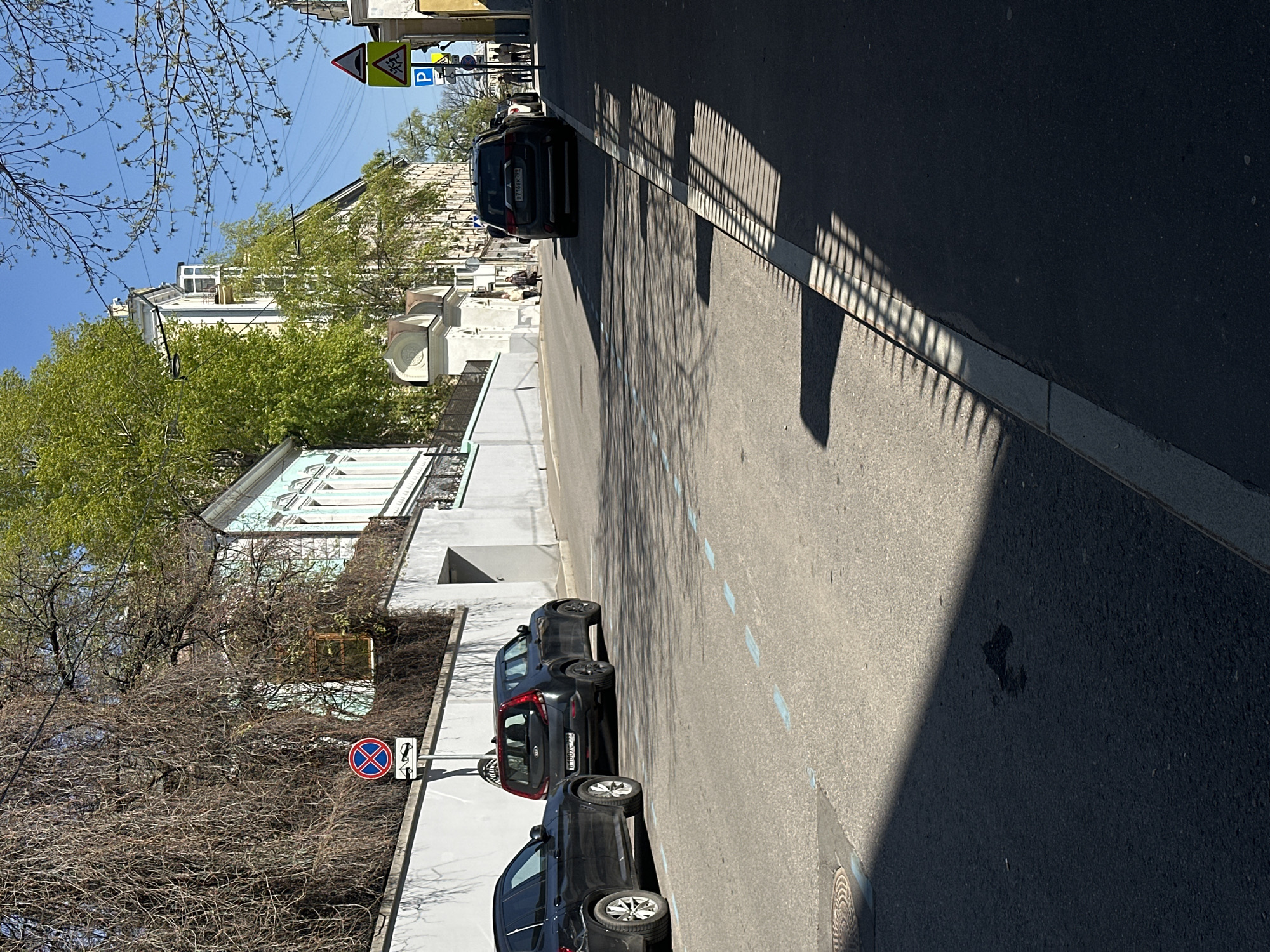 The image depicts a street scene in Moscow, Russia. Parked cars line the left side of the street near a building. A no-parking sign is visible next to the cars. A shadow from a fence is cast across the street. In the distance, more buildings are visible, along with traffic signs.