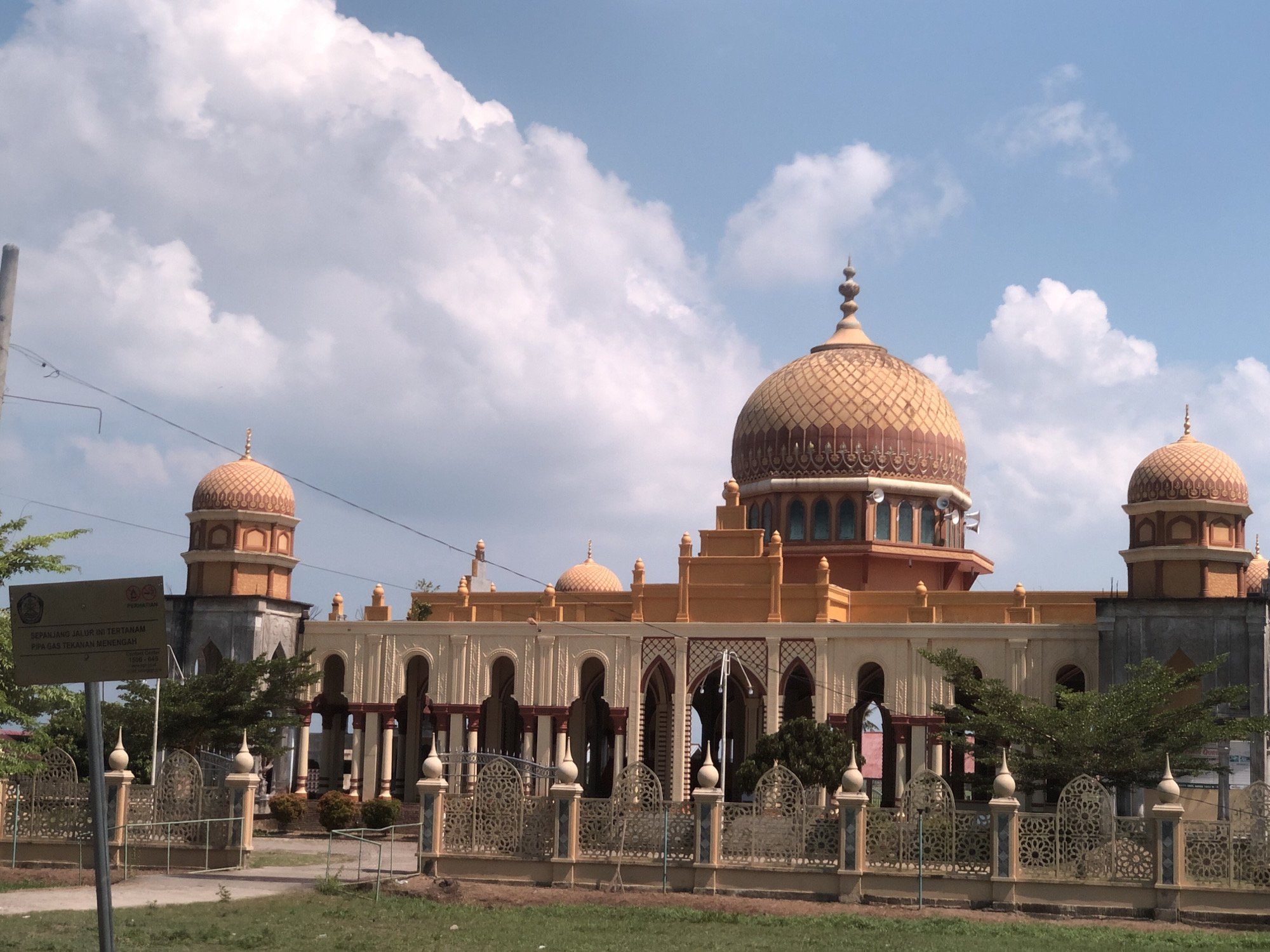 This is a photograph of a mosque located in the city of Peurupok, Indonesia. The mosque features multiple domes, with a prominent large dome in the center and smaller domes on either side. The architecture is characterized by arches, columns, and ornate fencing in the foreground. A signpost is visible to the left of the mosque, with text in Indonesian indicating a warning about underground gas pipes. The sky is blue with scattered white clouds. The surrounding environment appears to be grassy with some trees. There are no people or animals visible in the image.
