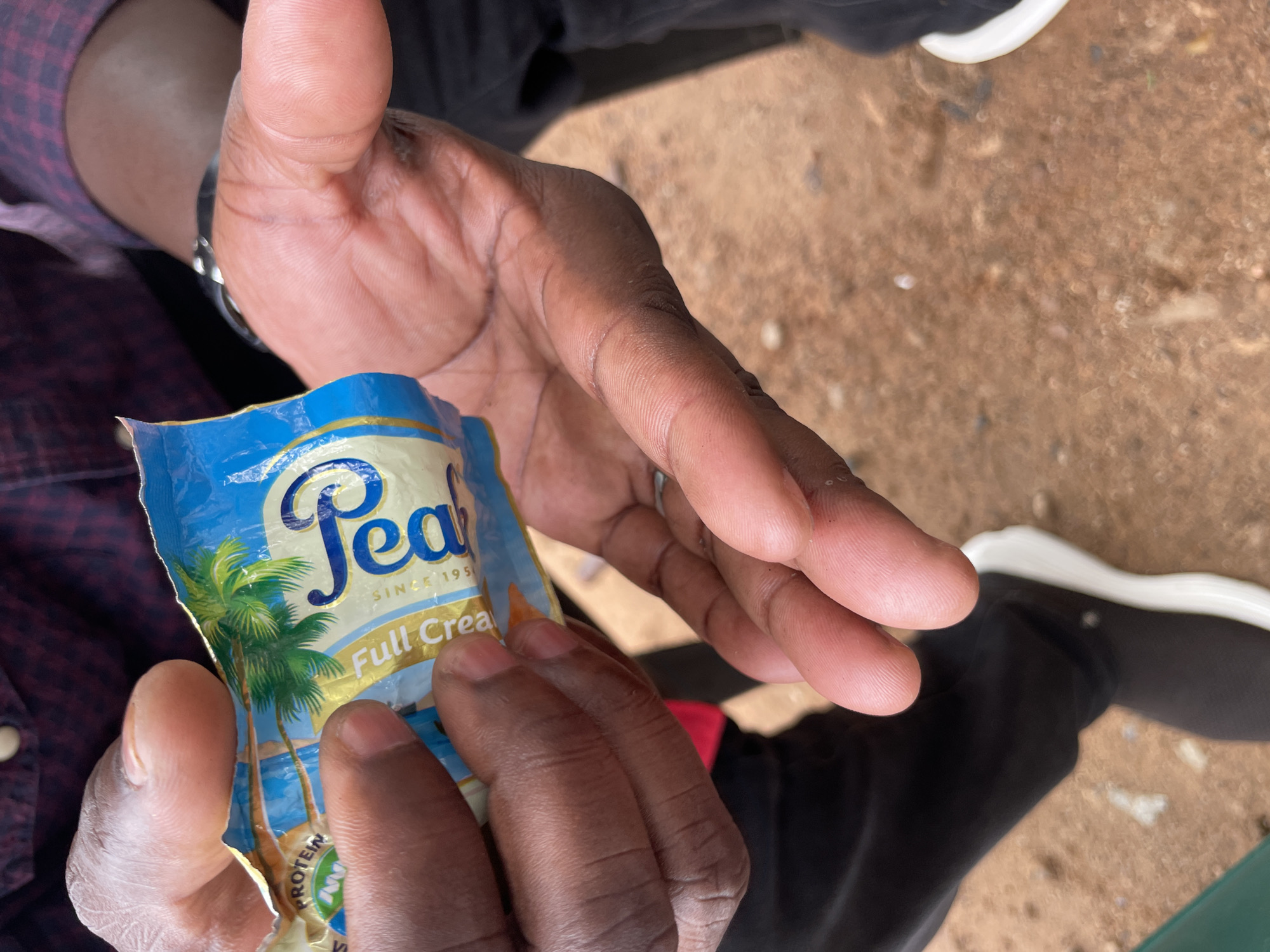 A close-up, eye-level shot captures a pair of dark-skinned hands manipulating a crumpled blue and gold packet. The packet, partially open, displays a palm tree graphic and text that includes "Peal" and "Full Cream". One hand holds the packet open, while the other appears to be reaching into it. The background is blurred but suggests an outdoor setting with reddish-brown ground, possibly dirt or gravel. The person's clothing, visible in the upper portion of the frame, is a dark, patterned shirt. The lower portion shows dark pants and a white-soled shoe. The scene is consistent with an individual in Jimeta, Nigeria, consuming a packaged food product.