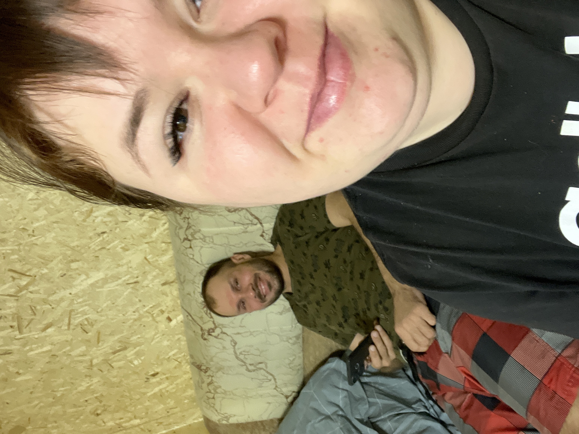 The image depicts two people in an indoor setting. A woman is positioned closer to the camera, her face filling a significant portion of the frame. She has short brown hair, fair skin, and is wearing a black t-shirt. Her expression is neutral with a slight smile. Behind her, a man is lying on a patterned cushion or mattress. He has a beard and is wearing a green patterned shirt. He appears to be smiling and looking at the camera. He is holding a black phone. The background includes what appears to be a wall with a wood-like paneling. The presence of bed-like or cushion-like furnishing indicates that this setting likely is a bedroom. There is insufficient visual information in the image to confirm the precise location in the city of Chaikovsky, Russia.