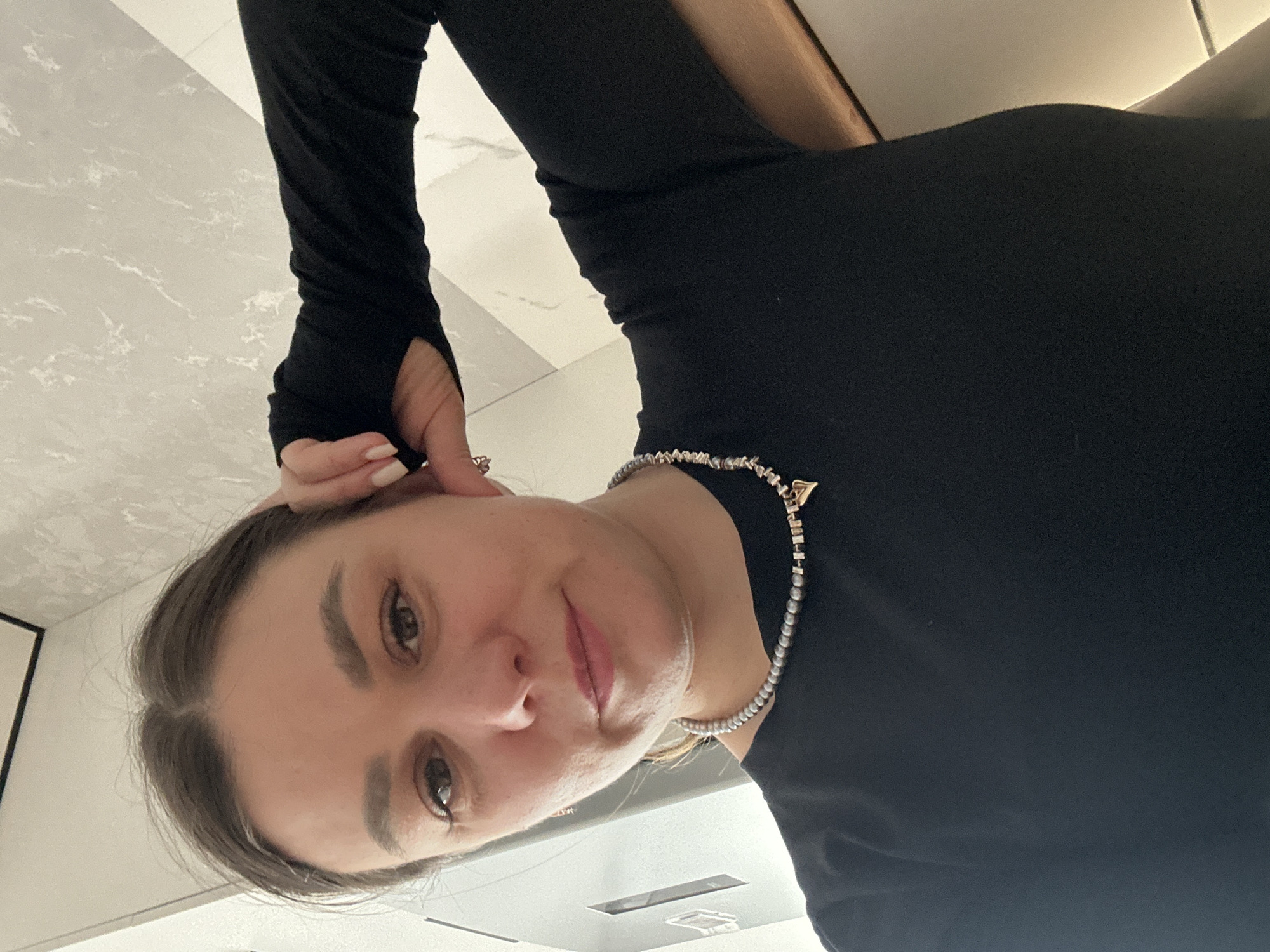 A woman is taking a selfie. She is wearing a black long-sleeved shirt with thumb holes and a beaded necklace with a small triangular charm. She is holding her hair near her ear. The background appears to be a room with light-colored tile flooring. There is no information to suggest the location is in Chaikovsky, Russia.