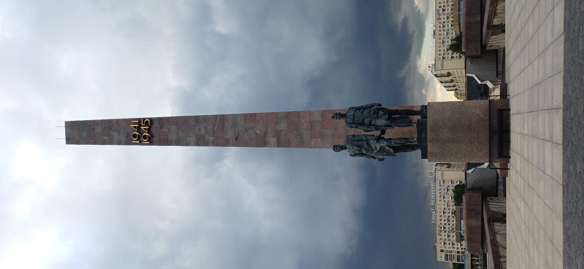 The image shows the monument to the Heroic Defenders of Leningrad, located in Victory Square, Saint Petersburg, Russia. A tall, imposing obelisk made of dark red granite stands in the center, inscribed with the years "1941" and "1945" in gold lettering. At the base of the obelisk are two bronze statues of Soviet soldiers, one holding a rifle. The monument is set against a dramatic sky with dark, storm clouds. In the background, apartment buildings and other structures typical of the city are visible, along with signs that read "SANKT-PETERBURG" and "GOROD GERØY LENINGRAD." The foreground features a paved plaza with large square tiles. The scene appears to be a solemn and overcast day.