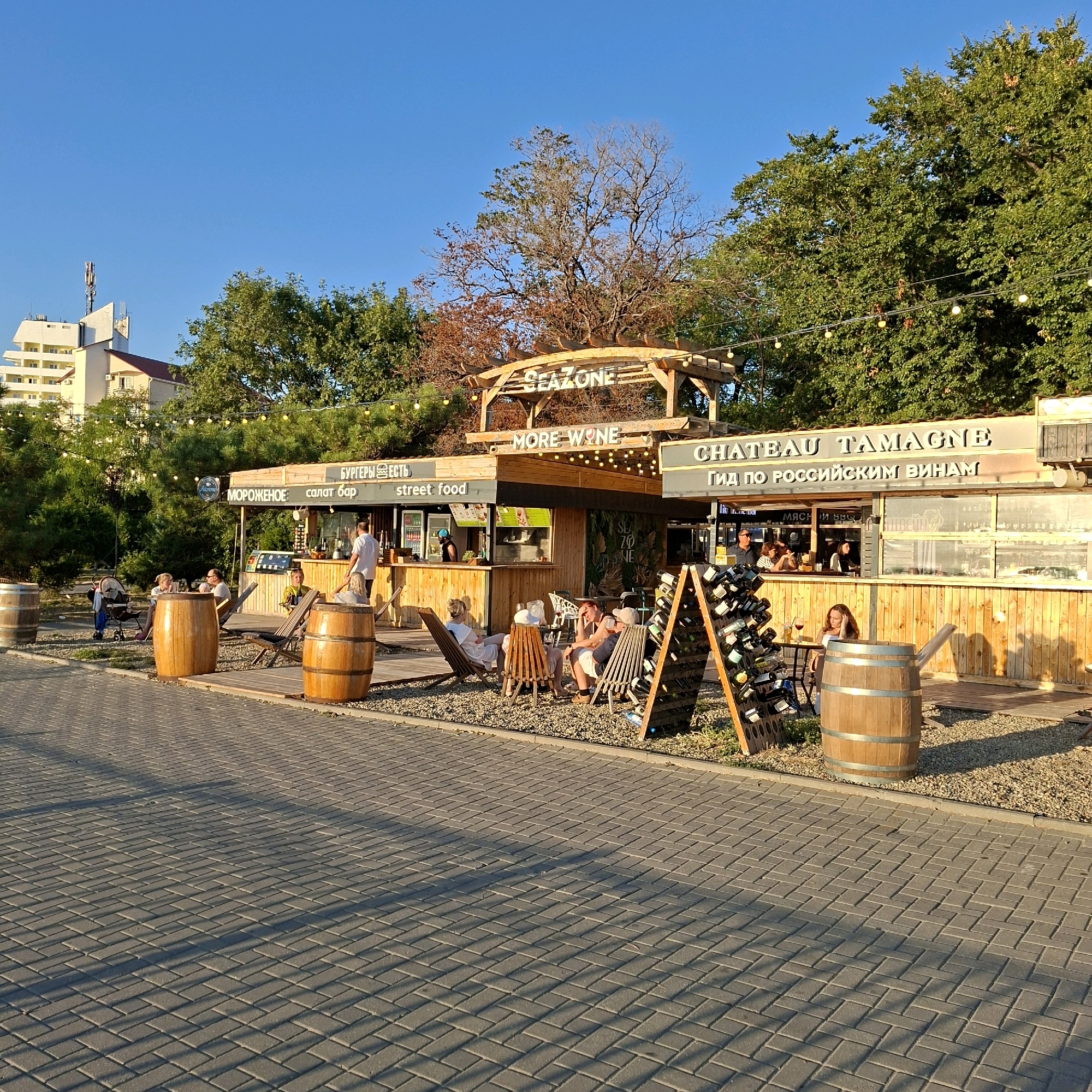 The image shows an outdoor seating area with a wooden structure and a bar in the background, likely a cafe or restaurant. The sky is clear and blue, and the sun is casting shadows on the brick-paved ground. Several people are seated at tables, enjoying drinks and conversation. A wine rack is visible on the right side of the image, filled with bottles. The overall atmosphere appears relaxed and social.