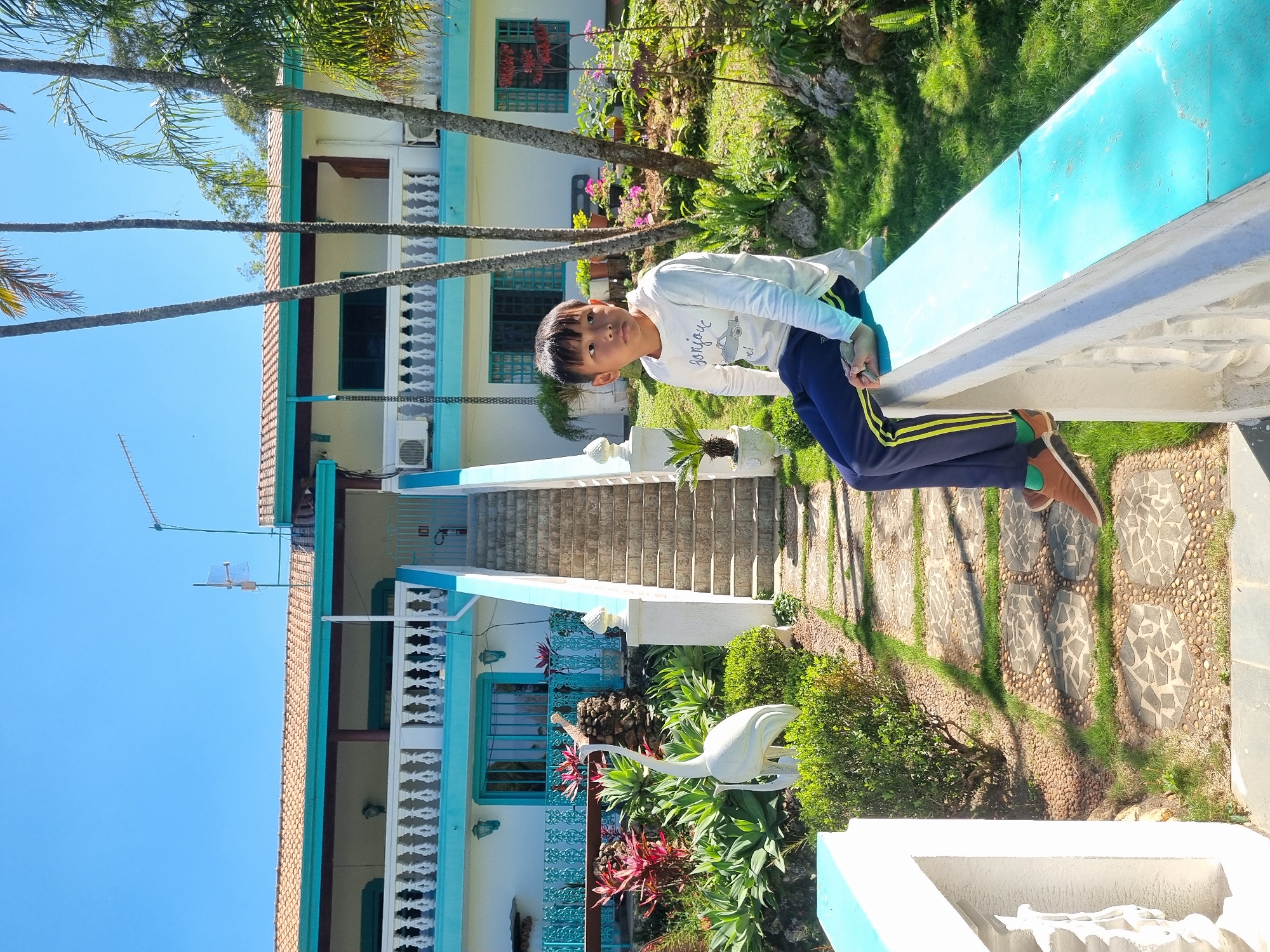 A young boy sits on a turquoise railing in front of a white building with a tiled roof and a balcony. The boy is wearing a white long-sleeved shirt and dark blue pants with yellow stripes. He is looking to his left. In the foreground, there is a stone pathway and lush greenery. A white statue of a bird stands in the garden. The sky is clear and blue. The building appears to be a hotel or guesthouse in Laranjal Paulista, Brazil, with its tropical foliage and distinct architectural style.